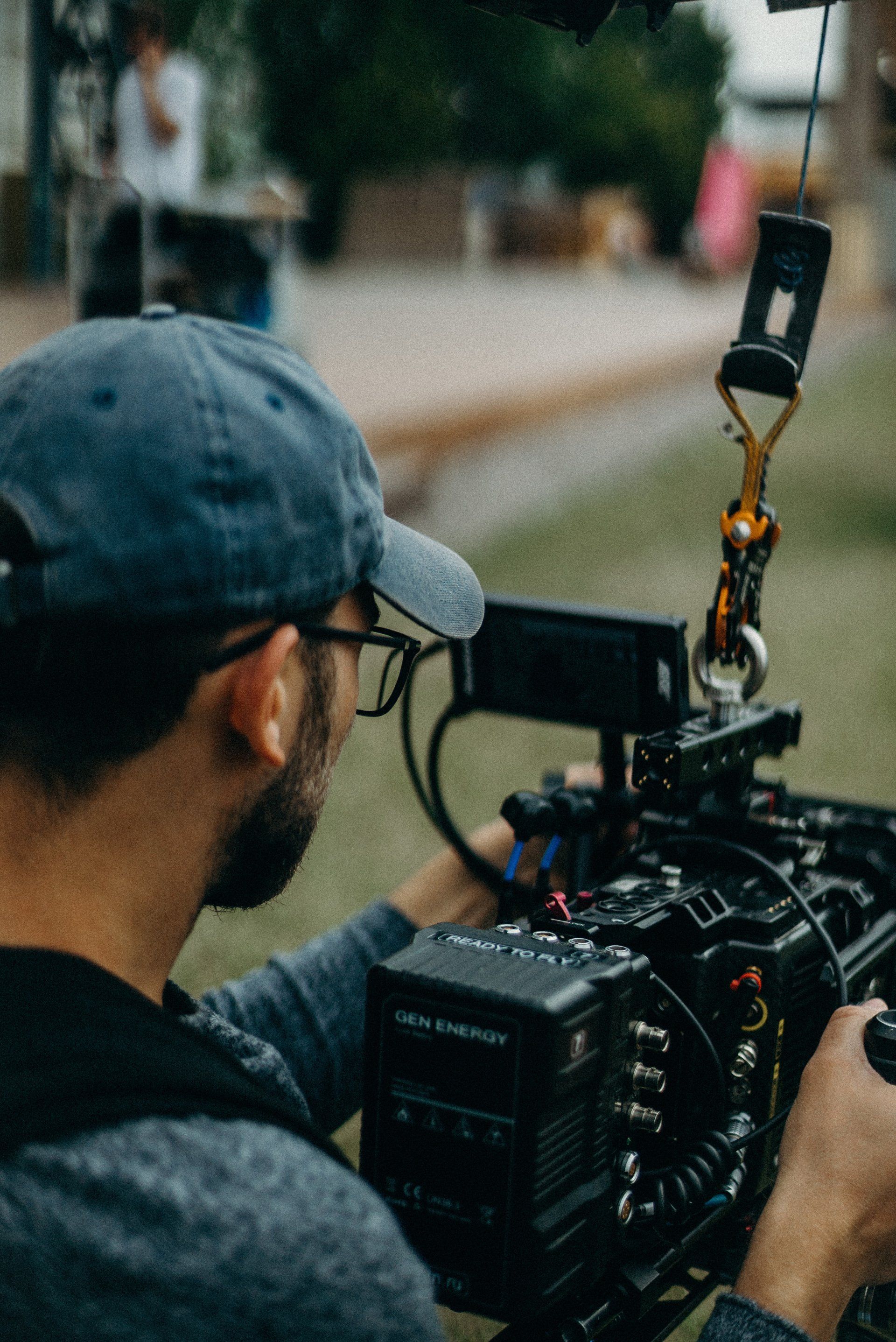 Person filming with a professional camera outdoors, wearing a blue cap and glasses.