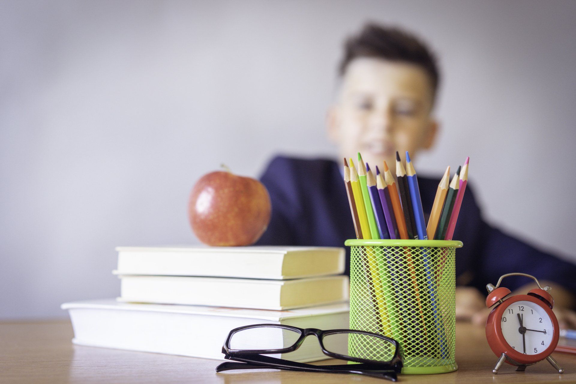 a boy is sitting at a desk with books , pencils , glasses and an apple .