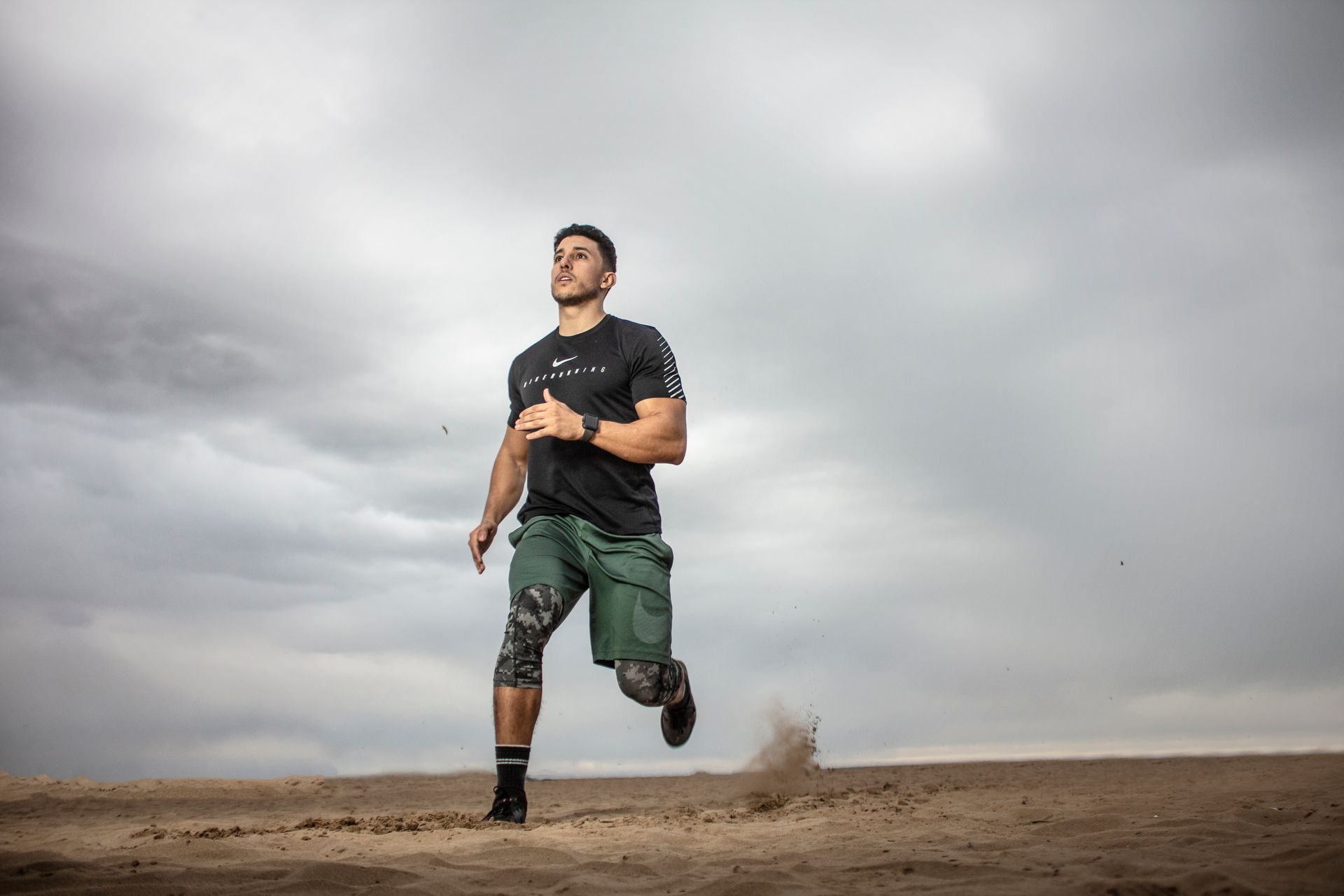 Man running on a sandy surface under a cloudy sky. He is wearing athletic wear and running shoes.