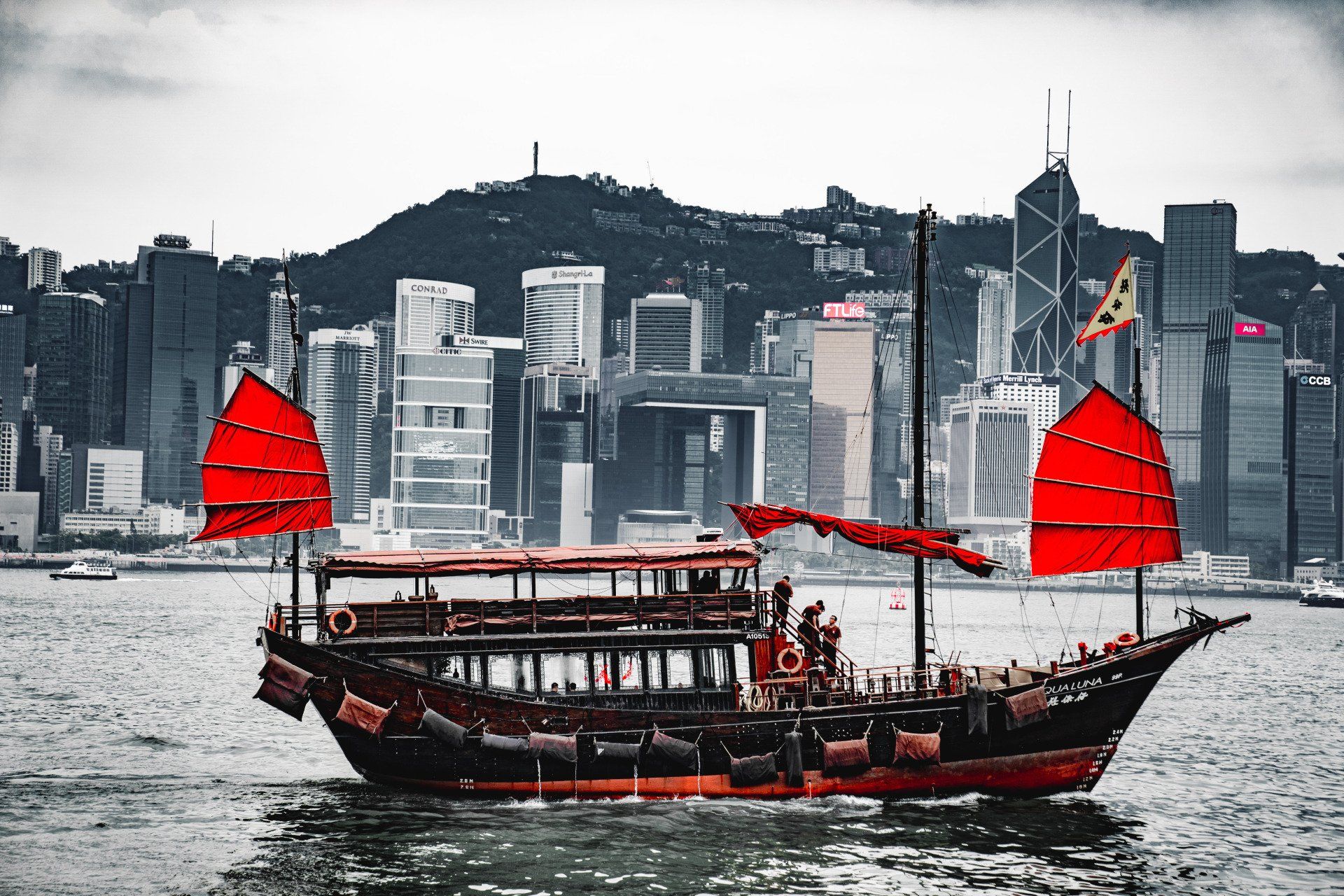 Traditional red-sailed junk boat on water, Hong Kong skyline in background.