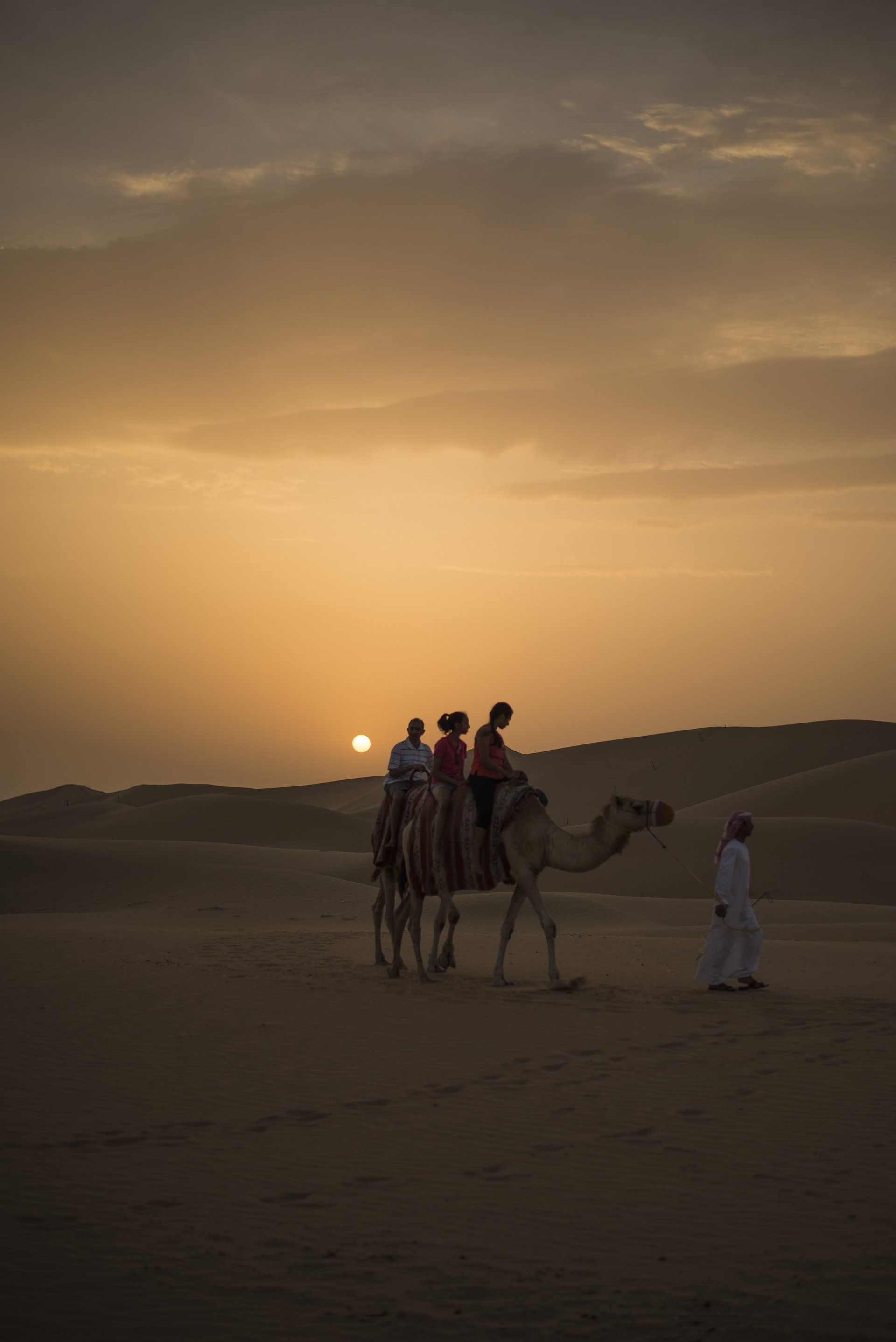 A group of people are riding camels in the desert at sunset.