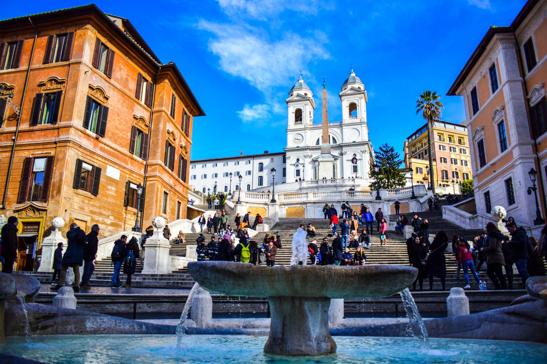 Scalinata di Trinità dei Monti a Roma, Italia, con persone che la salgono, fontana in primo piano e cielo azzurro.
