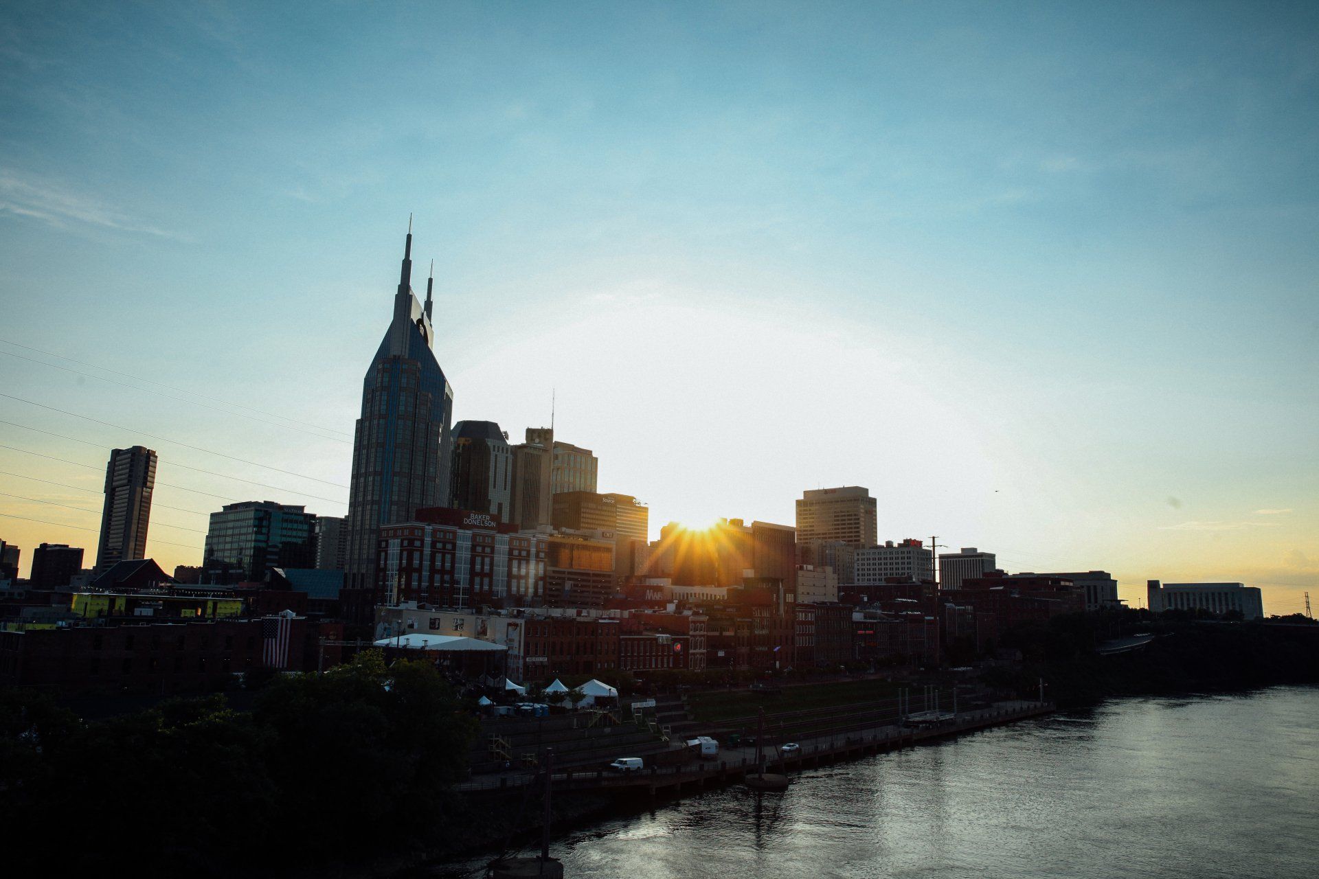 A city skyline with a river in the foreground and the sun shining through the clouds.