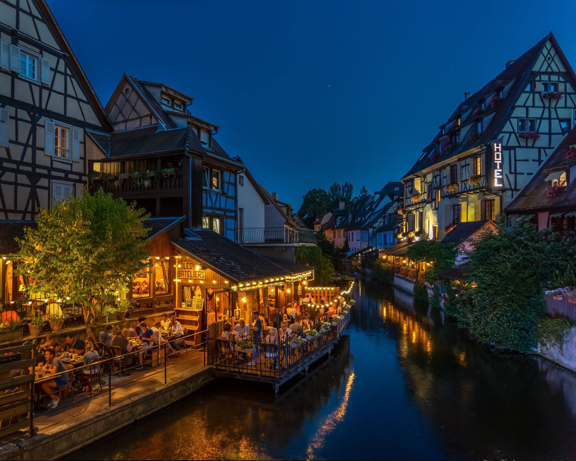 A group of people are sitting outside of a restaurant next to a river at night.