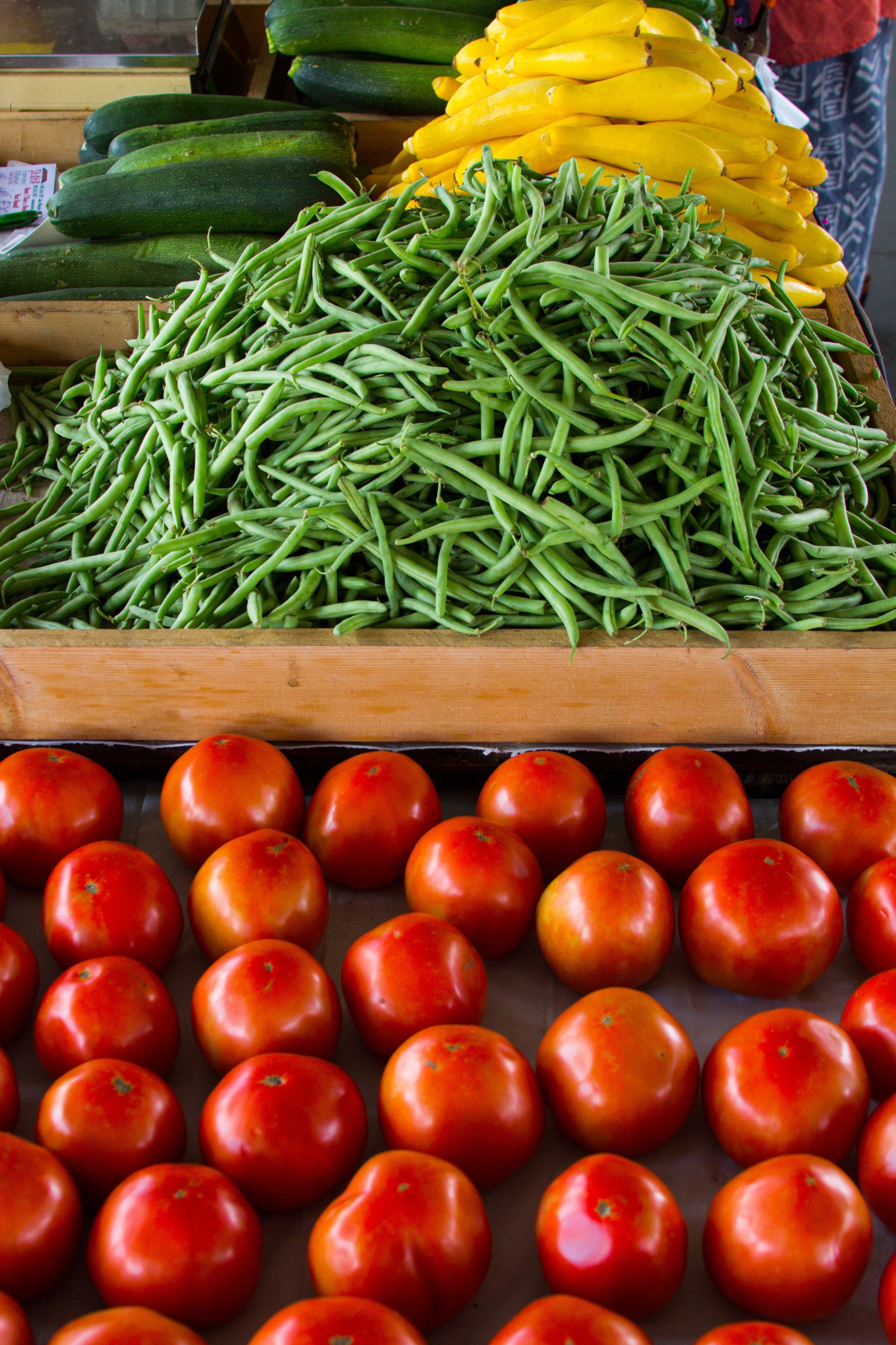 Fresh produce display at a farmer's market, featuring green beans, tomatoes, zucchini, and yellow squash.