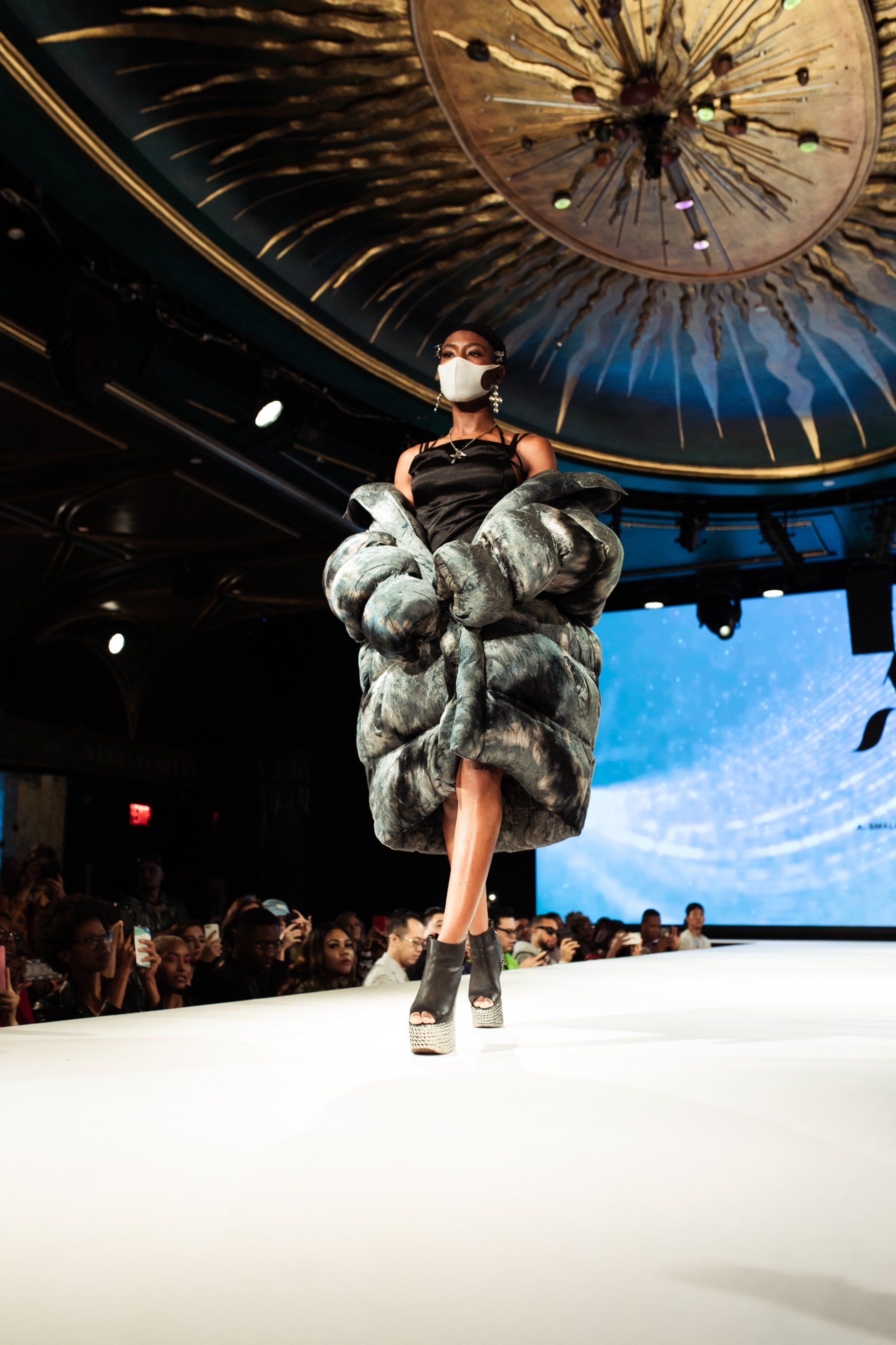 A model wearing a mask is walking down a runway at a fashion show.