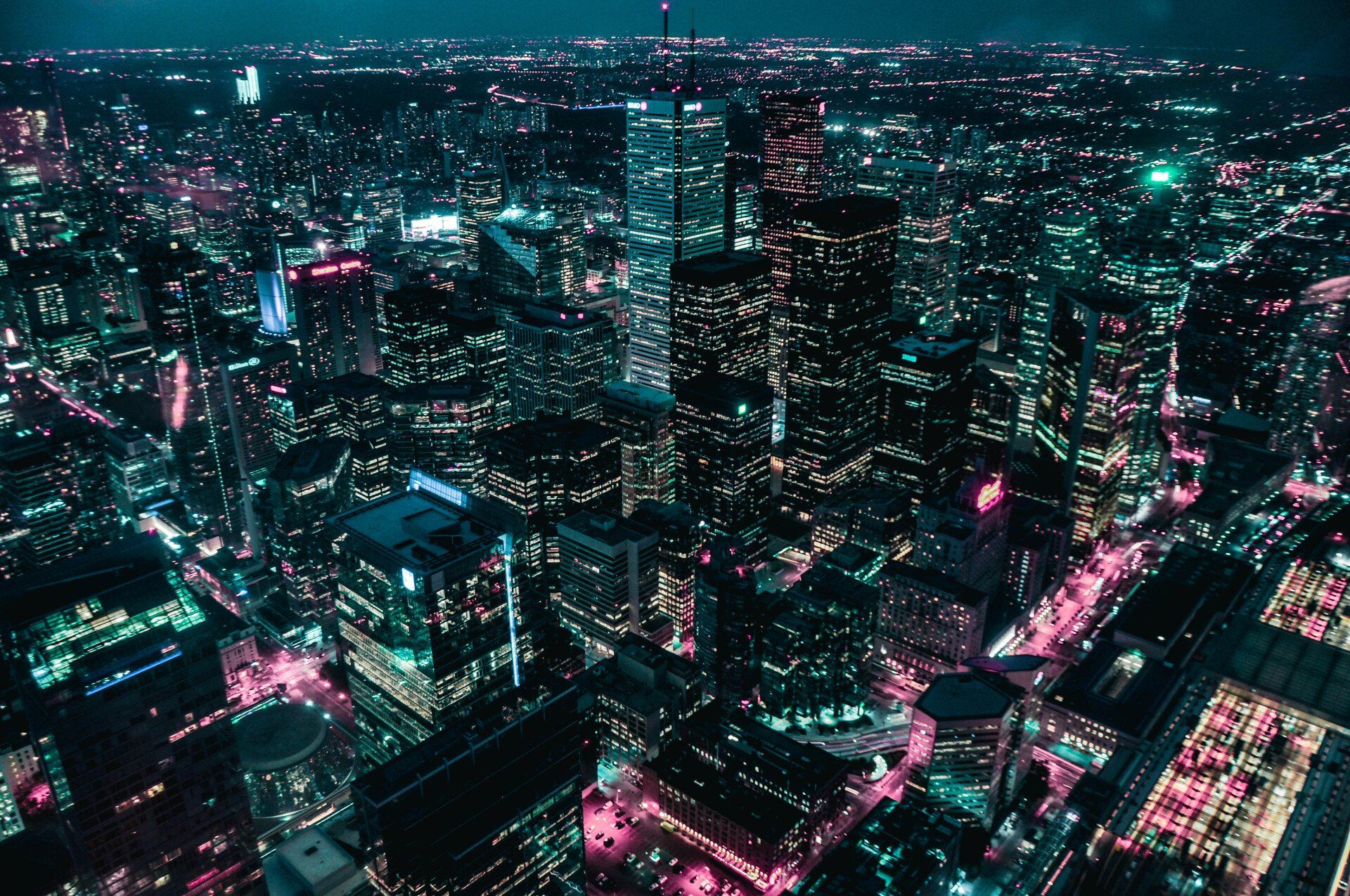 Aerial night view of a dense city skyline glowing with teal and pink lights.