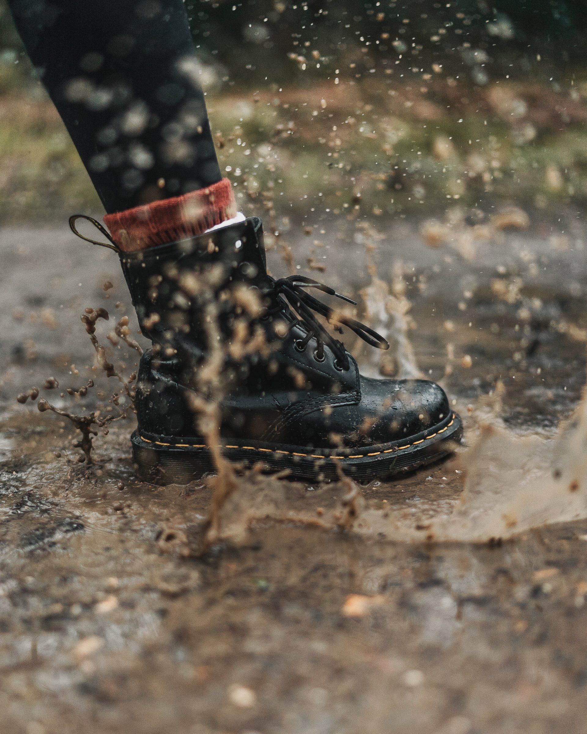 A black boot stepping into a muddy puddle, causing water and mud to splash around the footwear.