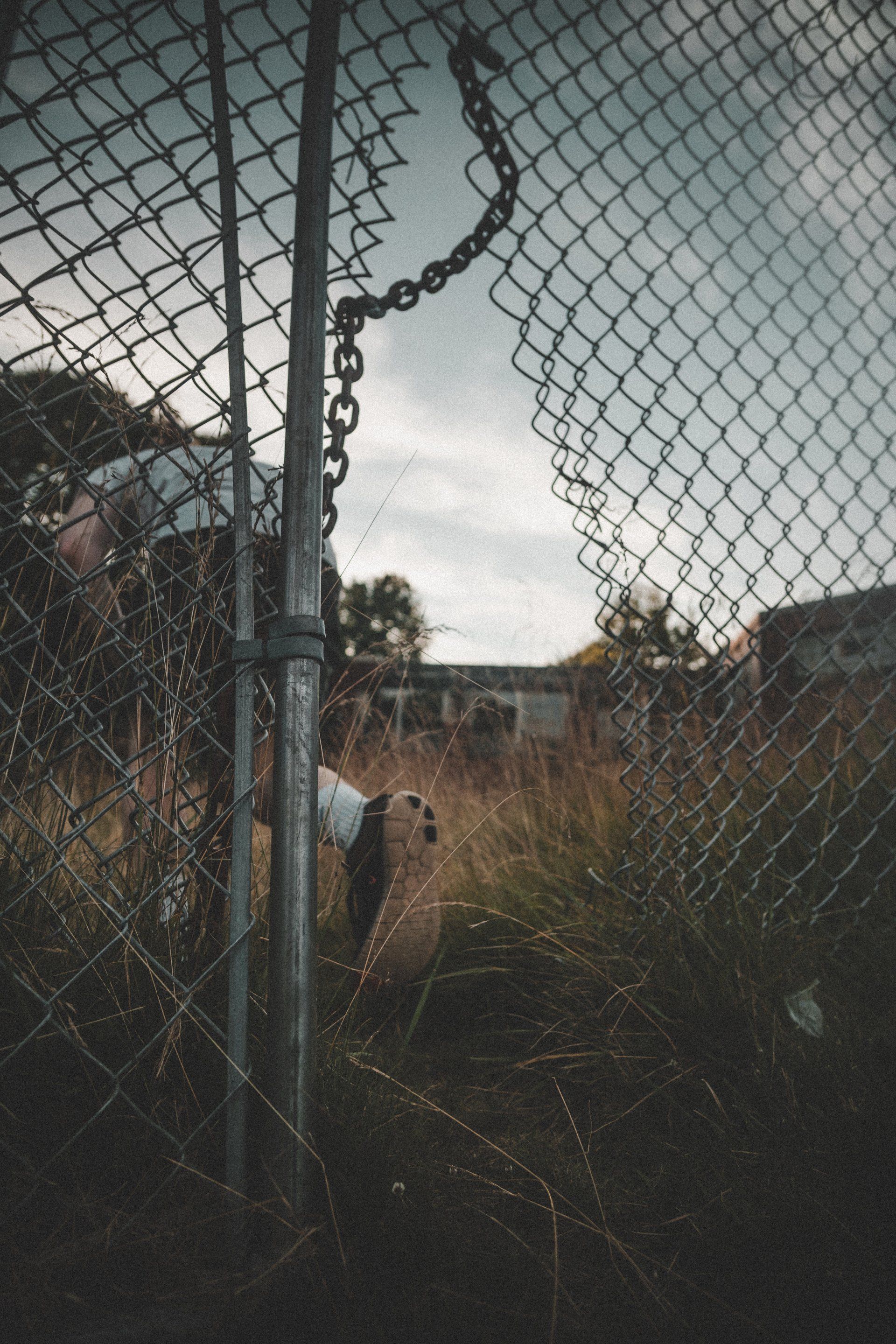 Breached chain-link fence, view into overgrown yard. Cloudy sky above.
