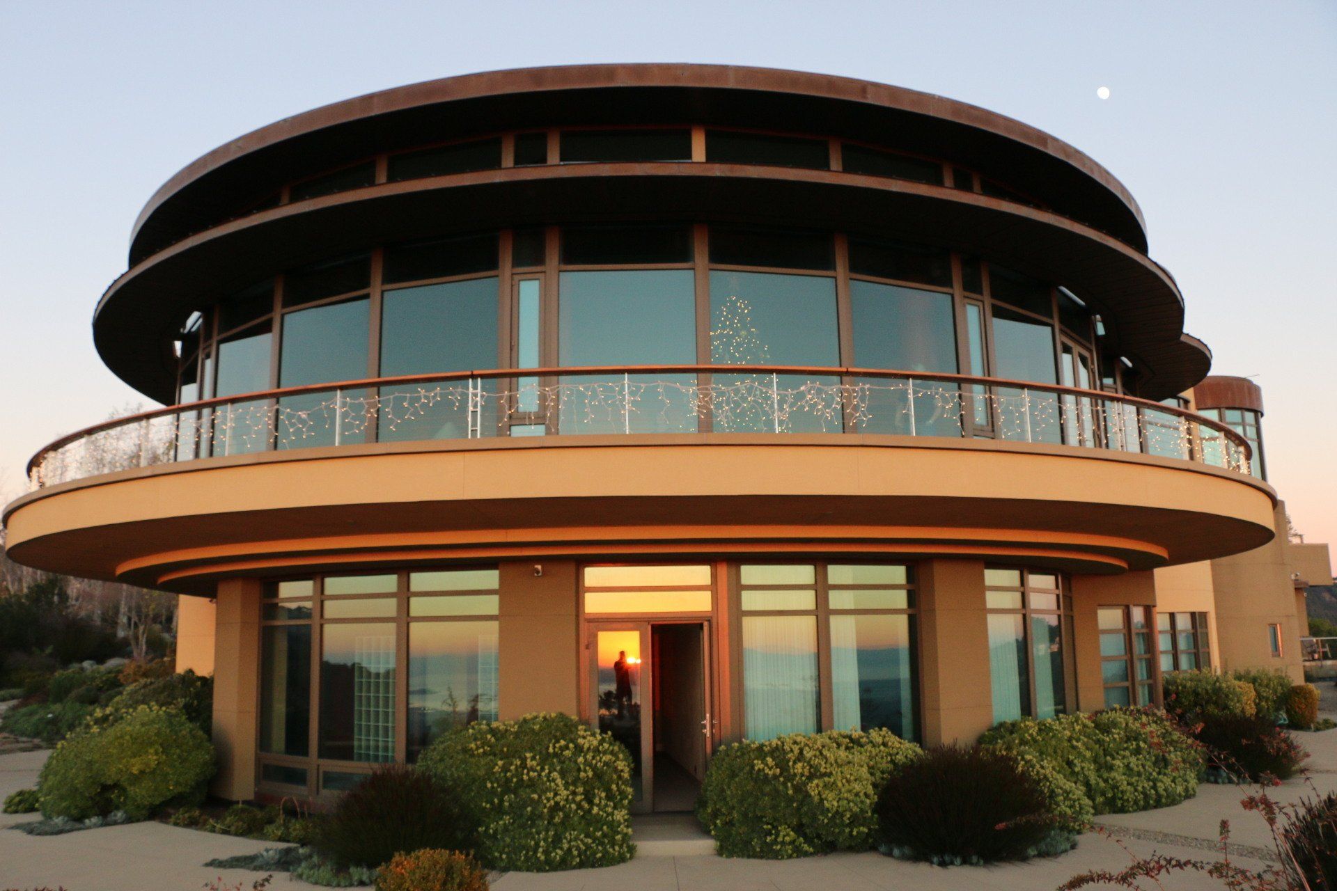 Casa moderna circolare con ampie finestre, balcone e finiture marroni al tramonto.