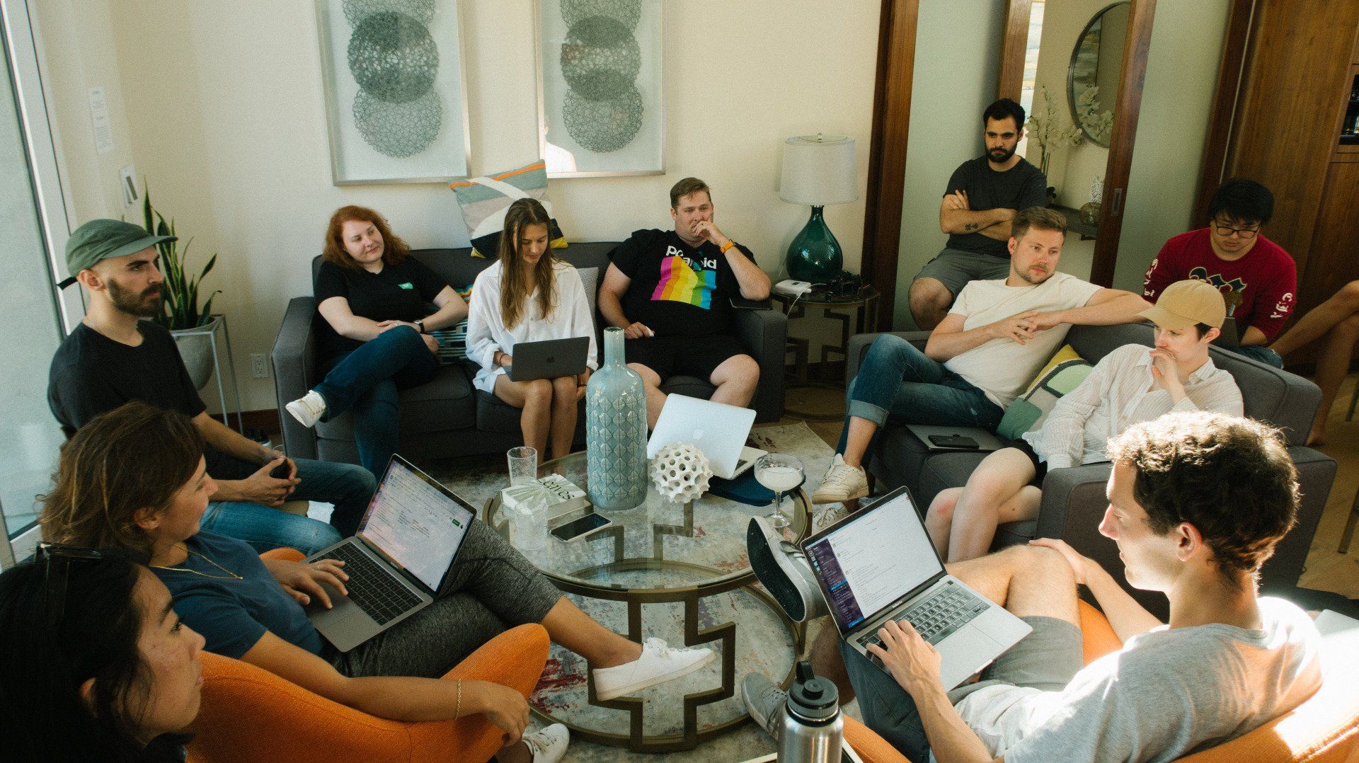 Group of people with laptops in a room, some sitting on couches, possibly in a meeting.