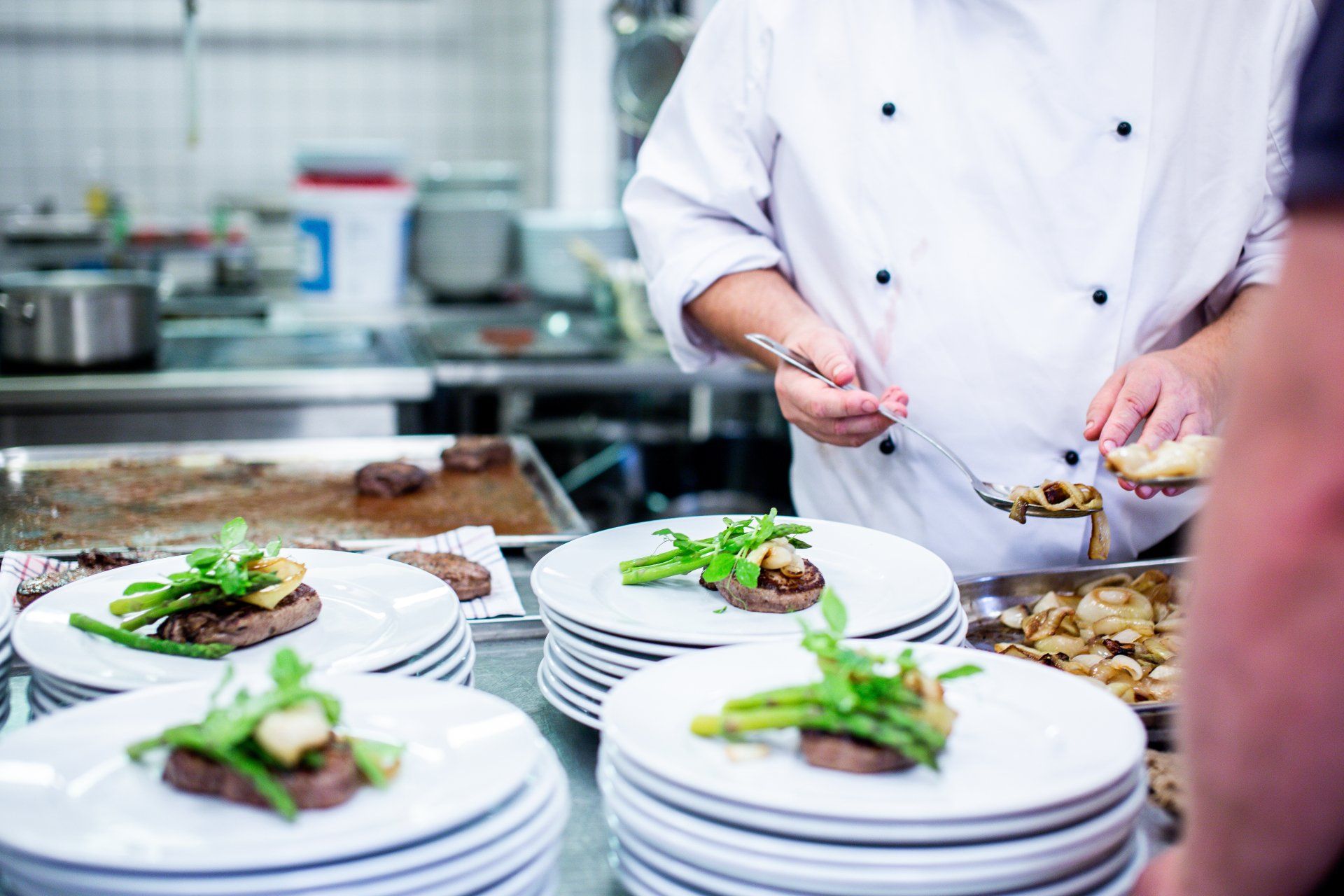 Chef plating food: steaks with asparagus and mushrooms on white plates in a commercial kitchen.