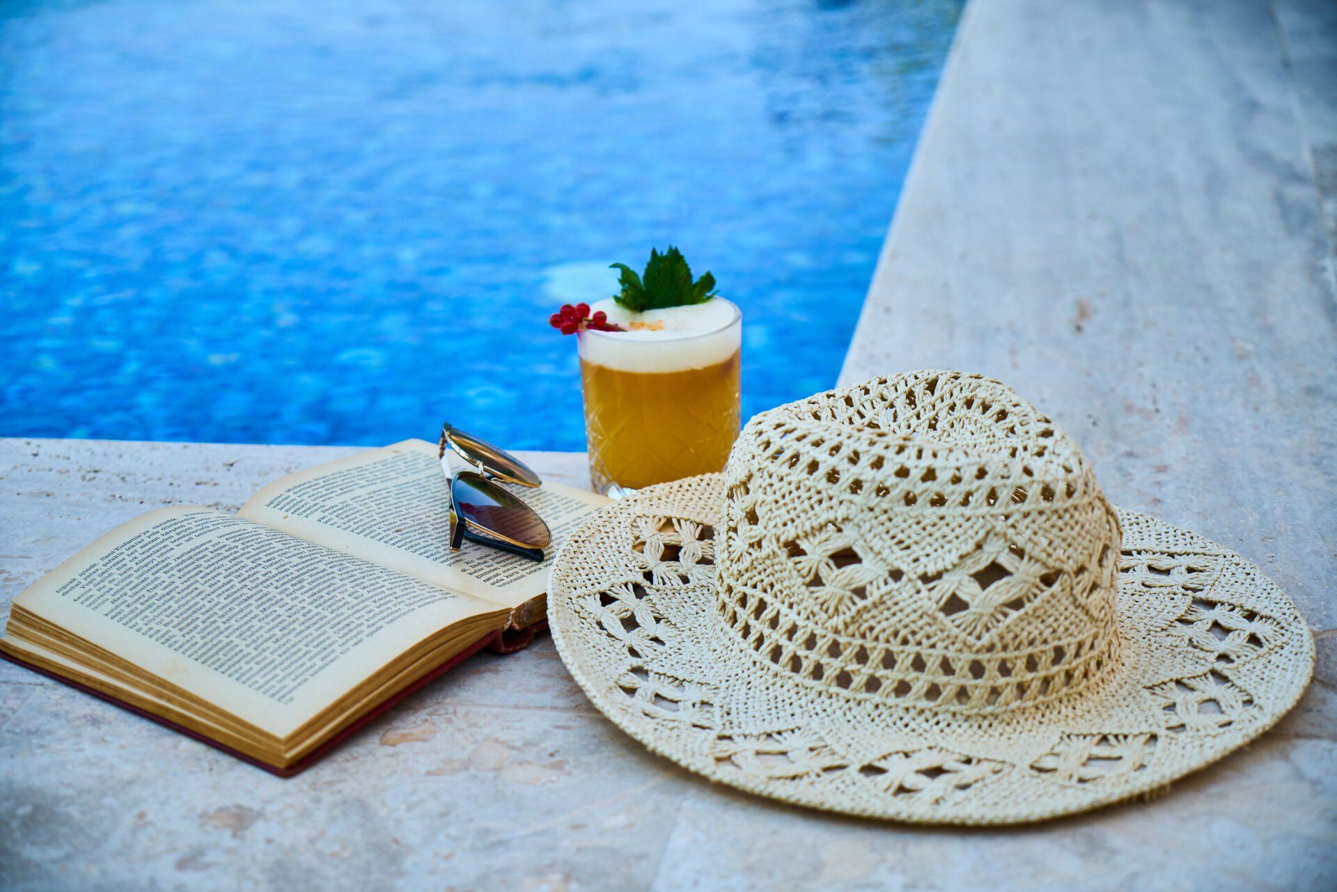 Livro aberto e chapéu de palha ao lado de uma bebida tropical à beira de uma piscina azul.