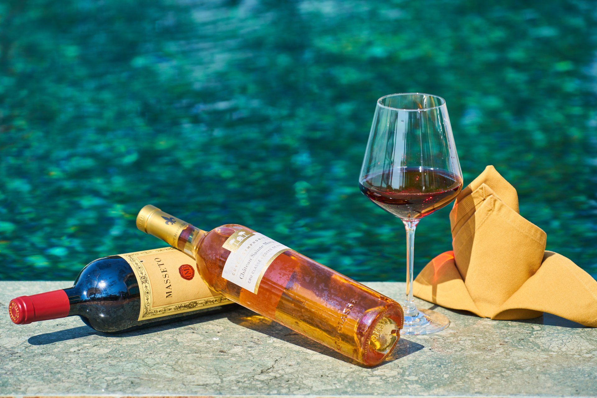 Wine bottles, a filled wine glass, and napkins on a sandy surface by turquoise water