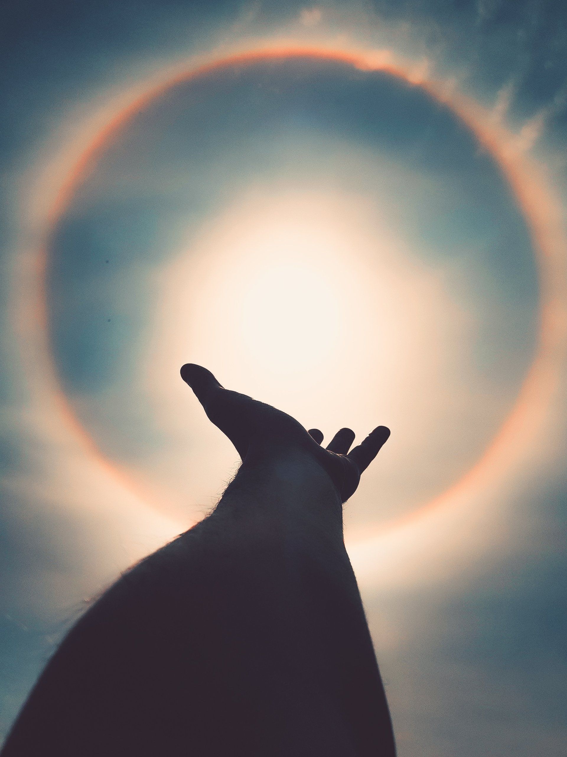 Silhouet van een hand die naar de zon reikt, omlijst door een regenboog aan een blauwe lucht.