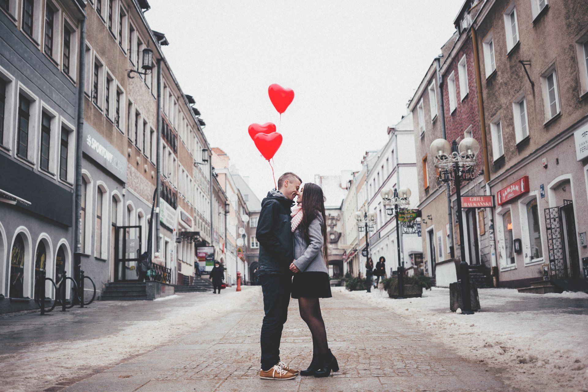 A man and a woman are kissing on a city street with balloons in the shape of hearts.