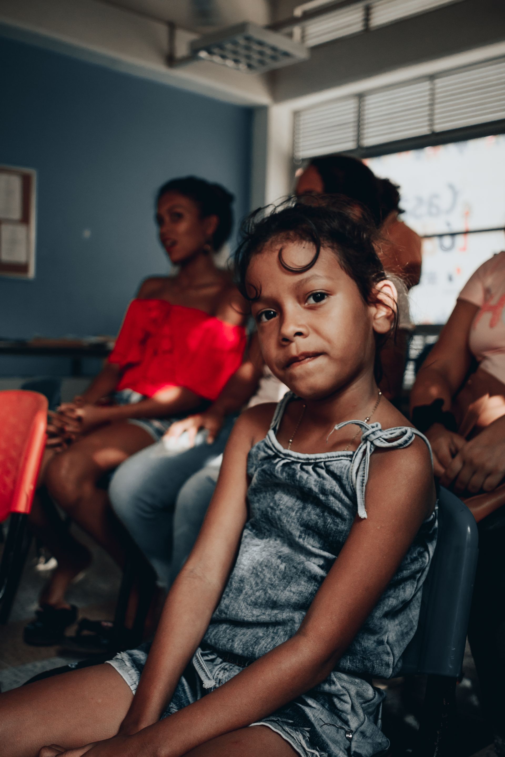 Comminty education, a little girl is sitting in a chair in a room with other people .