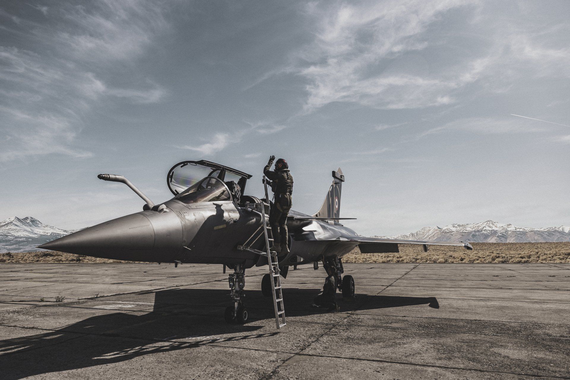 Pilot ascending a ladder to enter a gray fighter jet on an airfield, mountains in the background under a cloudy sky.