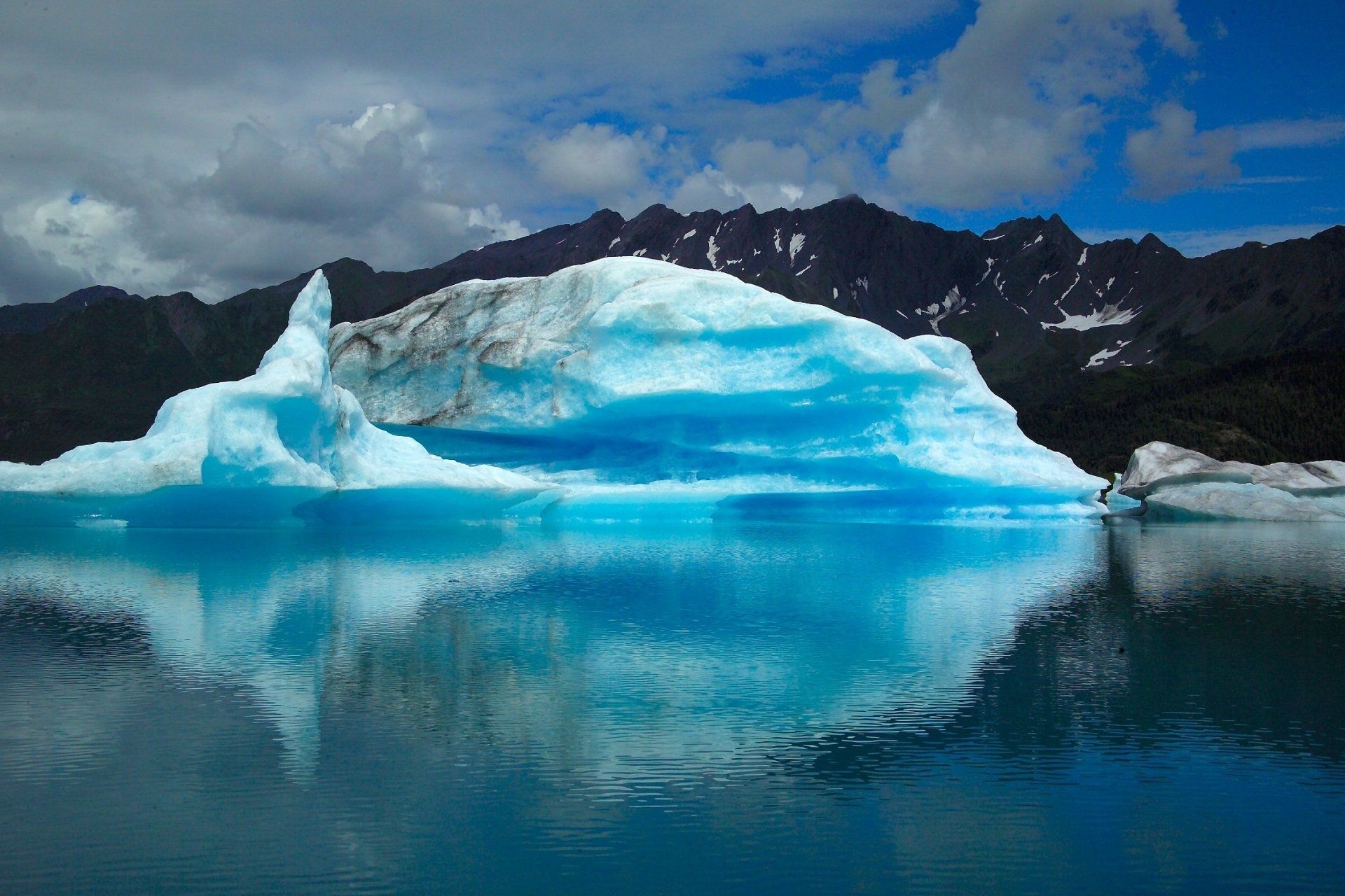A large blue iceberg is floating in a lake with mountains in the background.