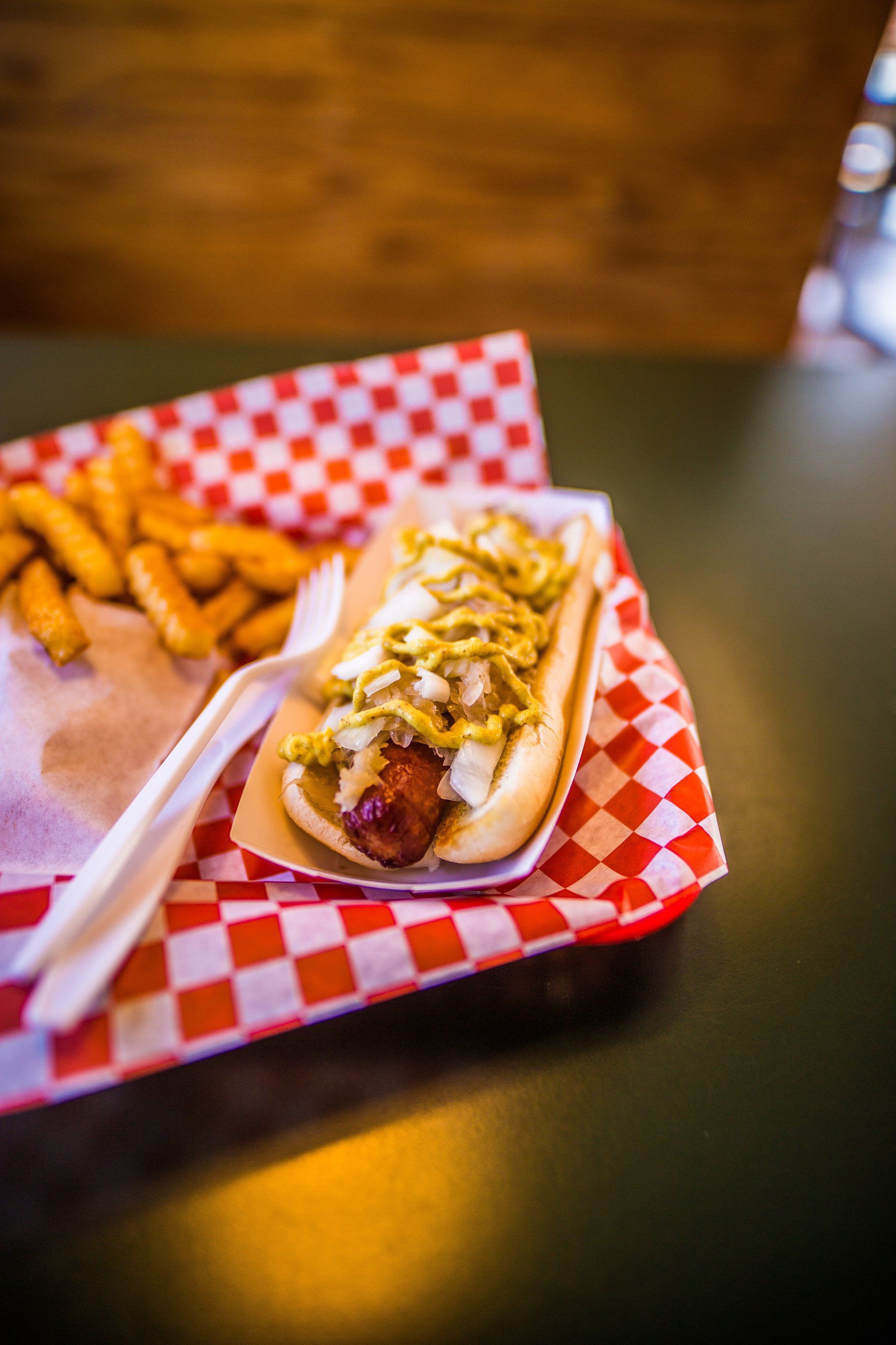 Hot dog with mustard and onions, fries, and a fork on a red and white checkered tray.