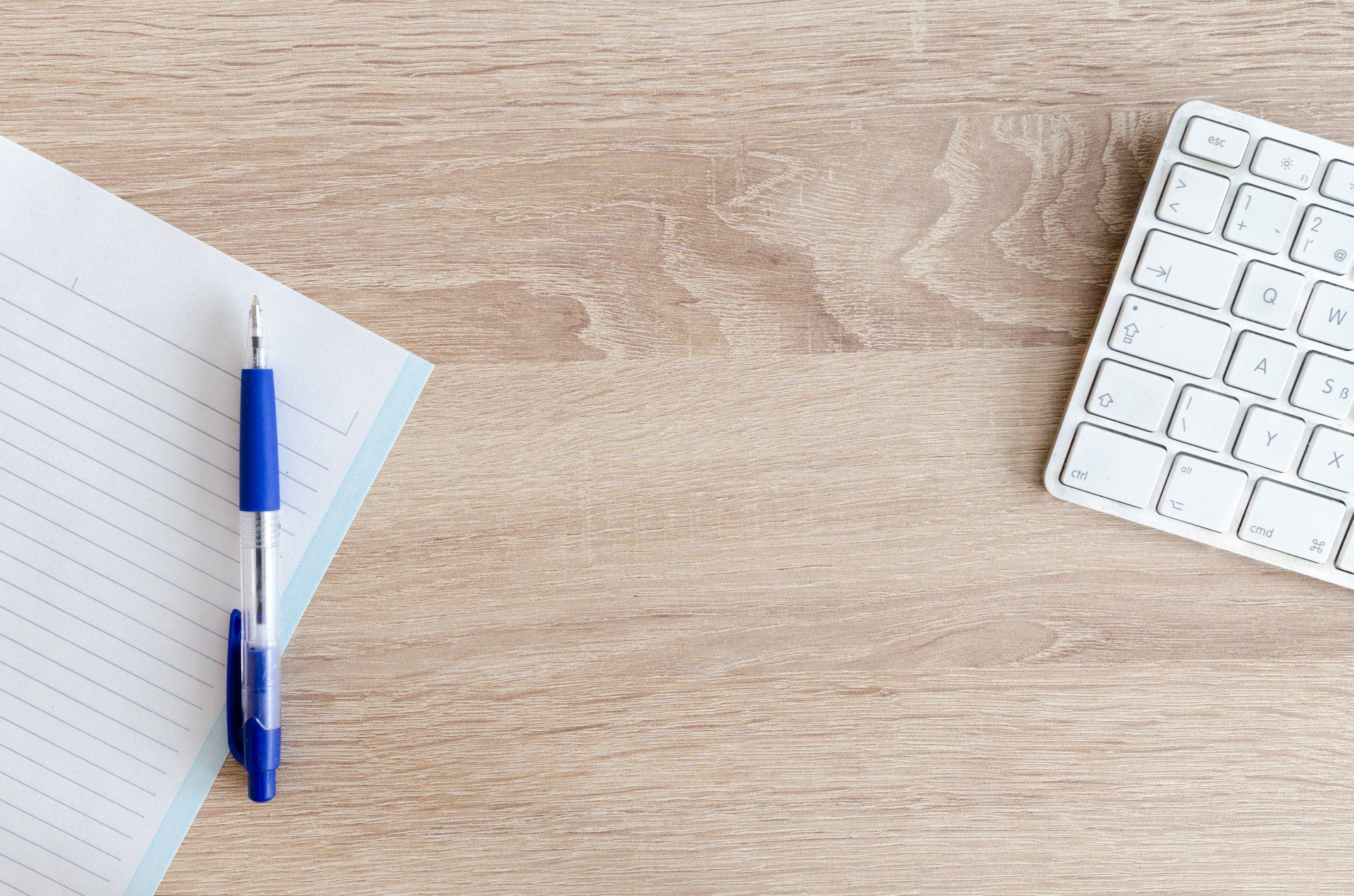 A pen , notebook and keyboard are on a wooden table.