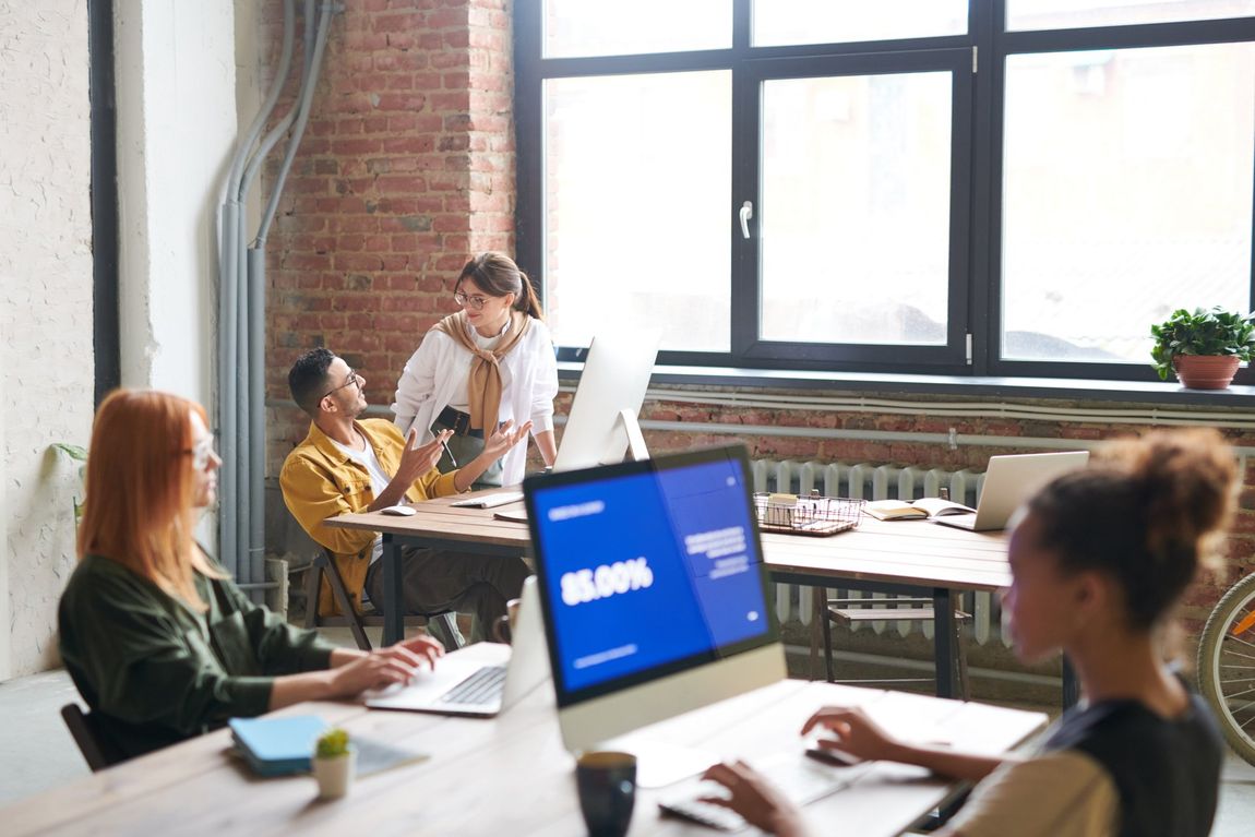 Group of diverse colleagues collaborating in a modern brick-walled office; working on laptops, discussing data.