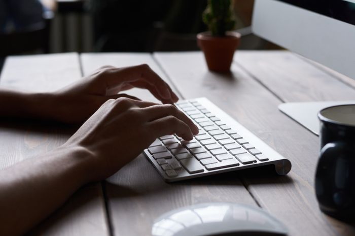 Hands typing on a silver keyboard at a wooden desk with a cactus and coffee mug nearby.