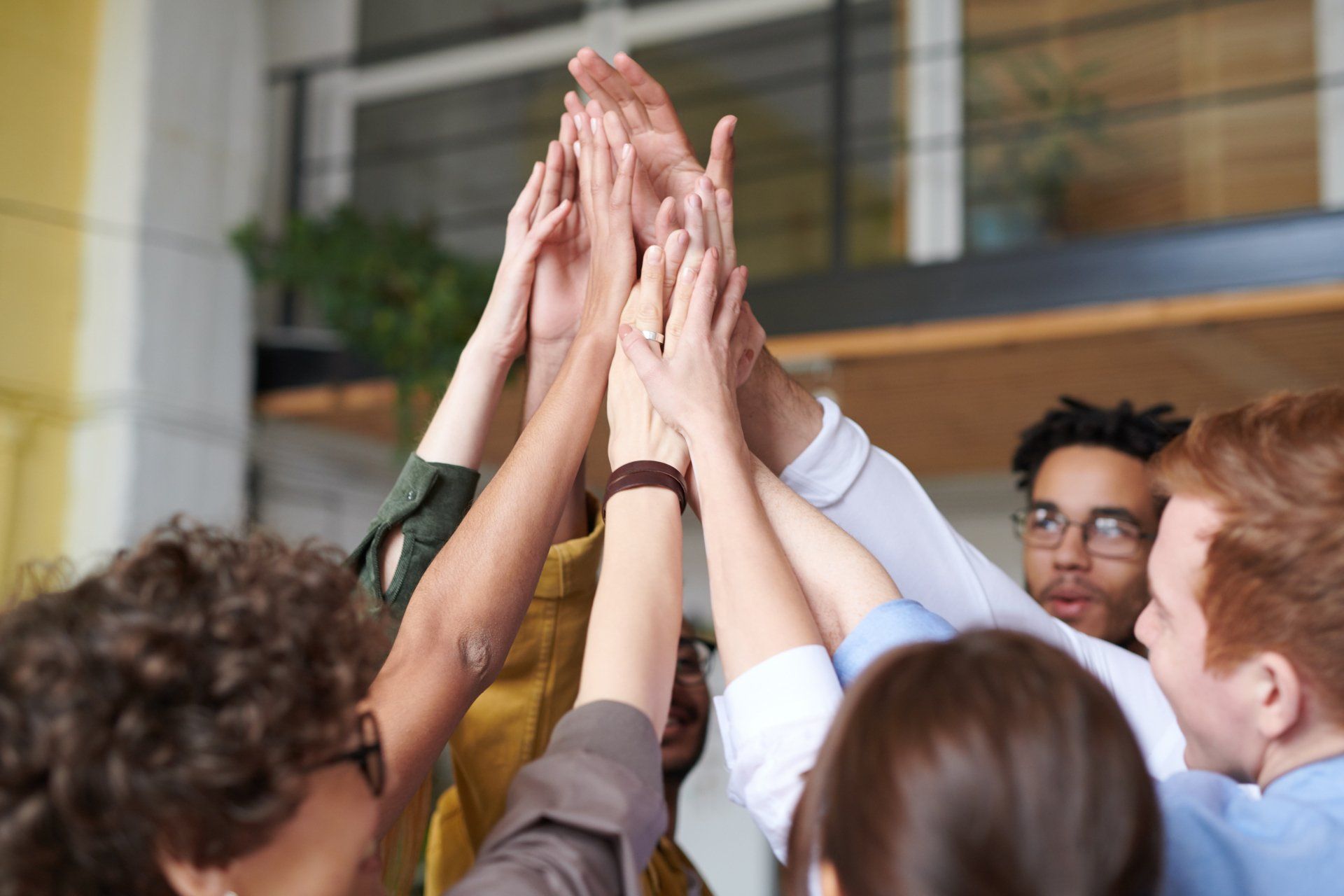 A diverse group of colleagues in an office setting reaching upward with their hands together in a celebratory high-five.