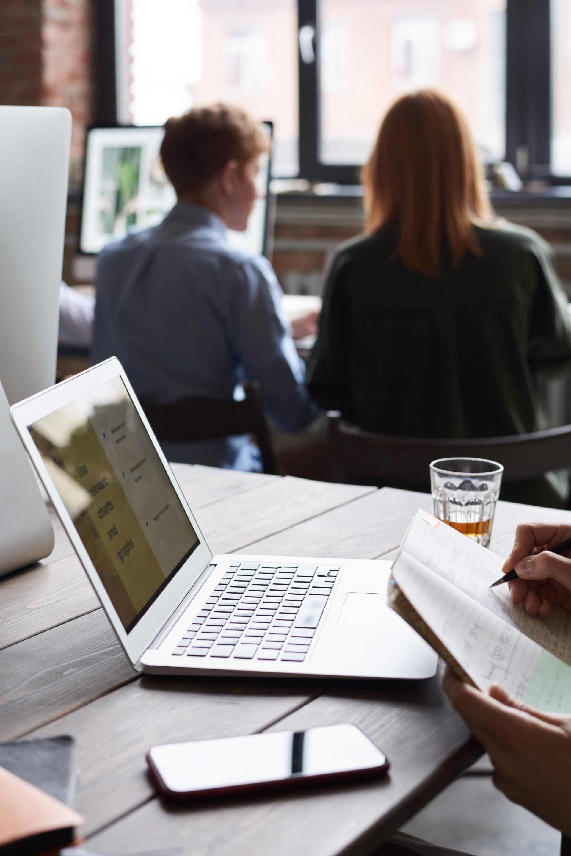 Laptop on desk, two people in background working, person taking notes.