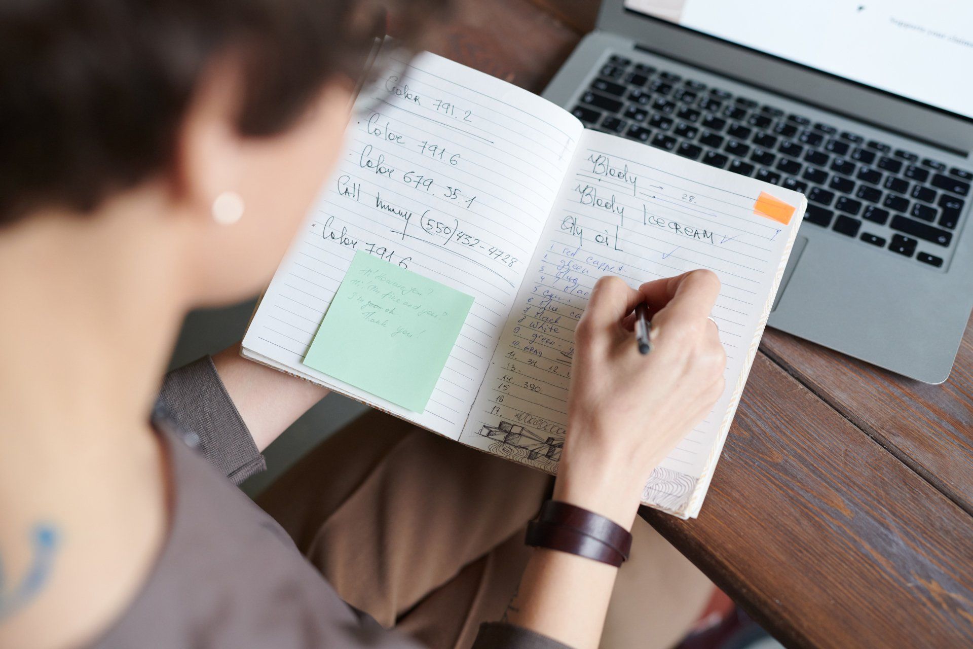A woman is writing in a notebook in front of a laptop.