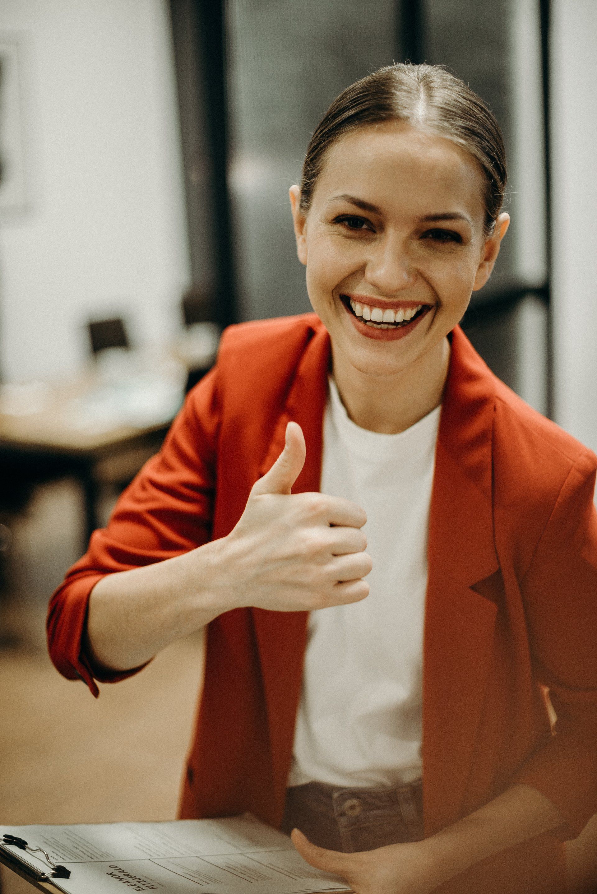 Woman in red blazer smiles and gives a thumbs-up in an office setting.