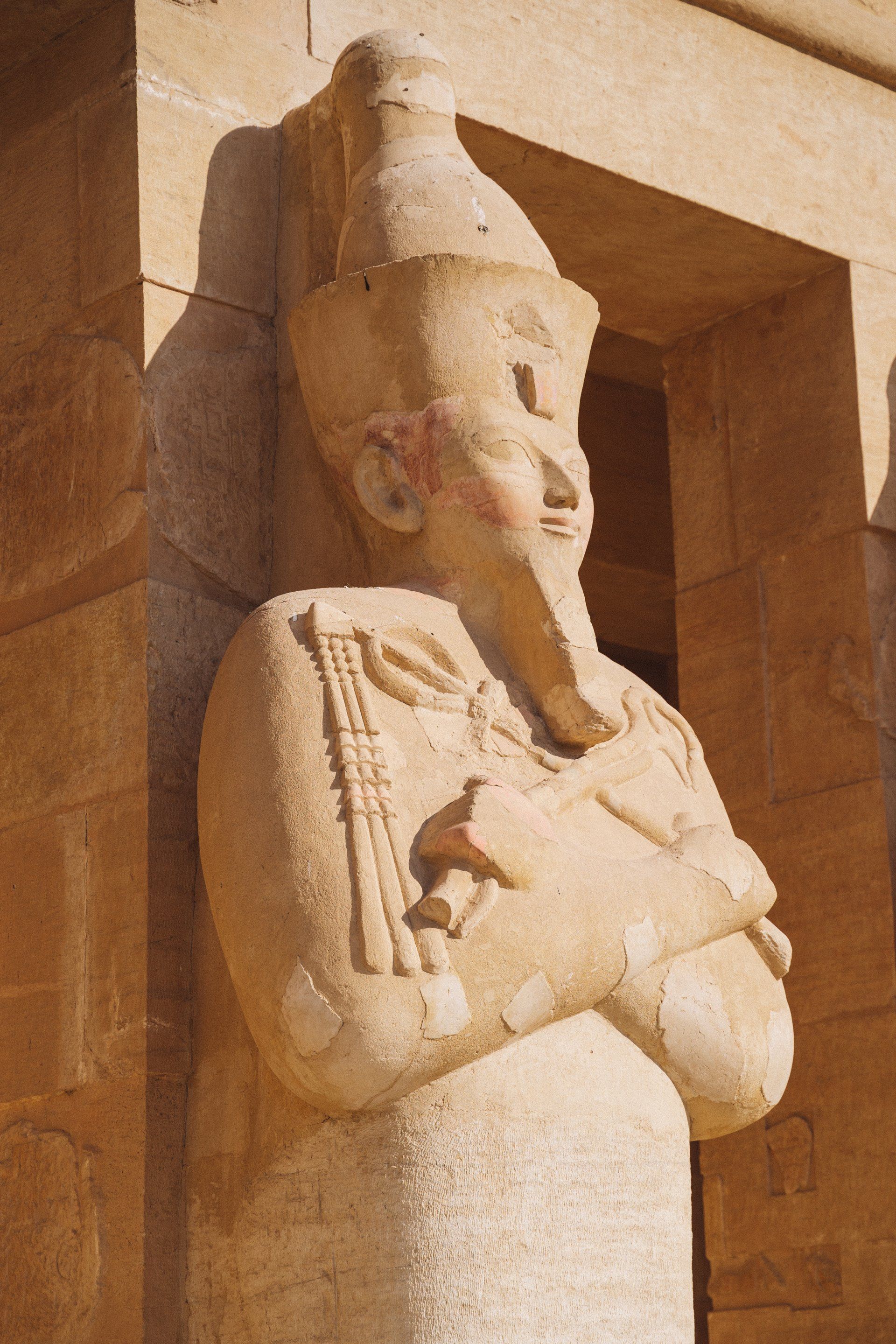 Stone statue of pharaoh in traditional headdress, arms crossed, at a temple.