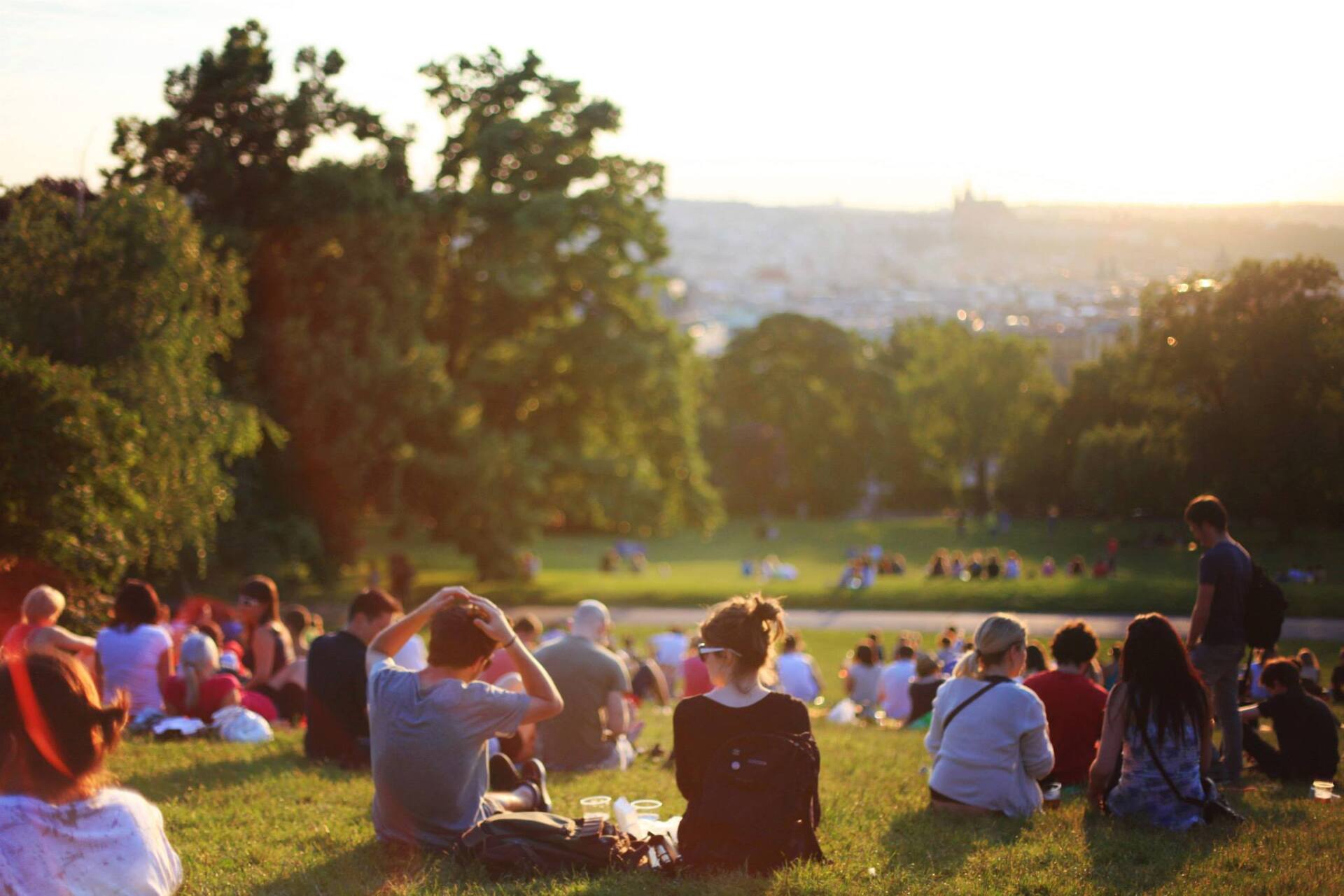 A crowd of people sits on a grassy hill overlooking a hazy city skyline during sunset.