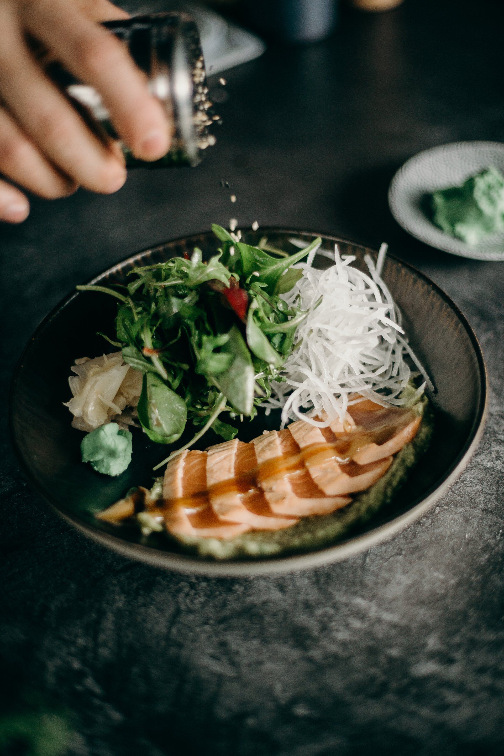 Persona che cosparge di semi di sesamo il sashimi di salmone con insalata e daikon.