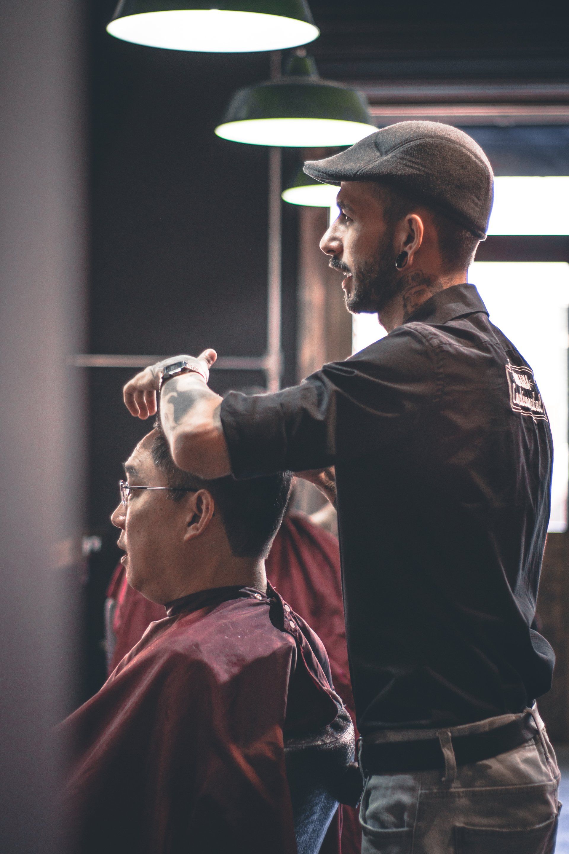Barbero cortando el pelo a un cliente en un salón bajo luces cenitales. Lleva gorra y tiene tatuajes.