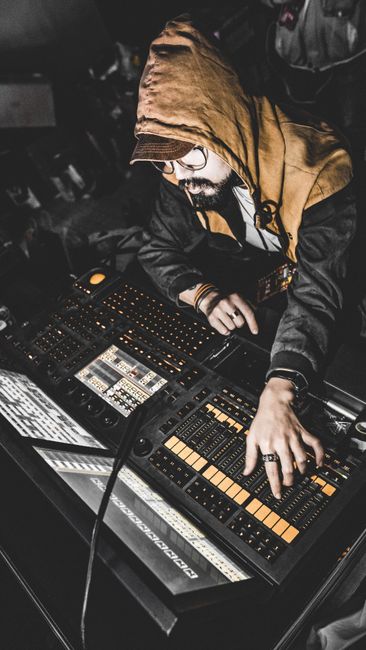 A man in a hoodie is playing a keyboard in a dark room with a sophisticated mixer