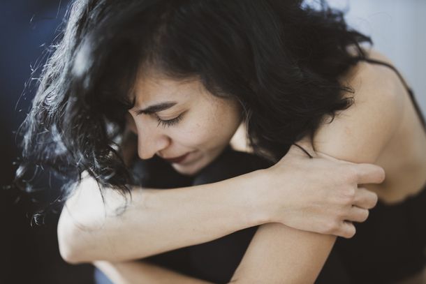 Woman with dark hair, eyes closed, embraces herself with a sad expression, indoors.