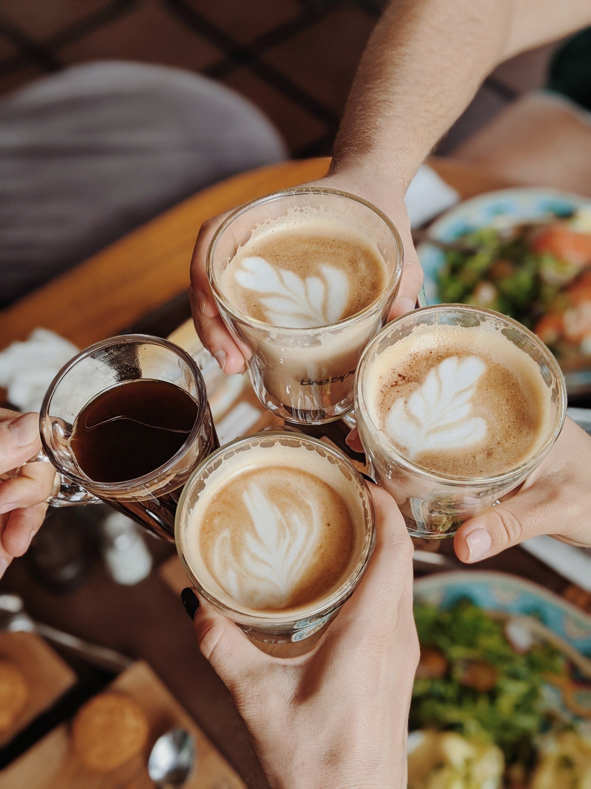 Four hands holding coffee cups, toasting. Two lattes, one black coffee. Blurred background of food.