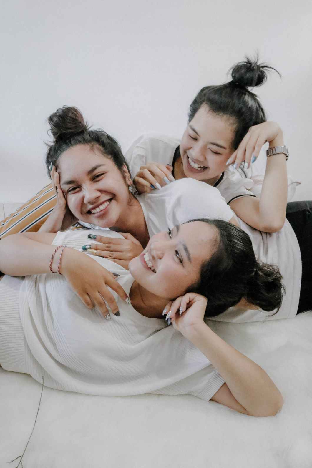 Three young women in white shirts, smiling and laughing, lying on a white fluffy surface.