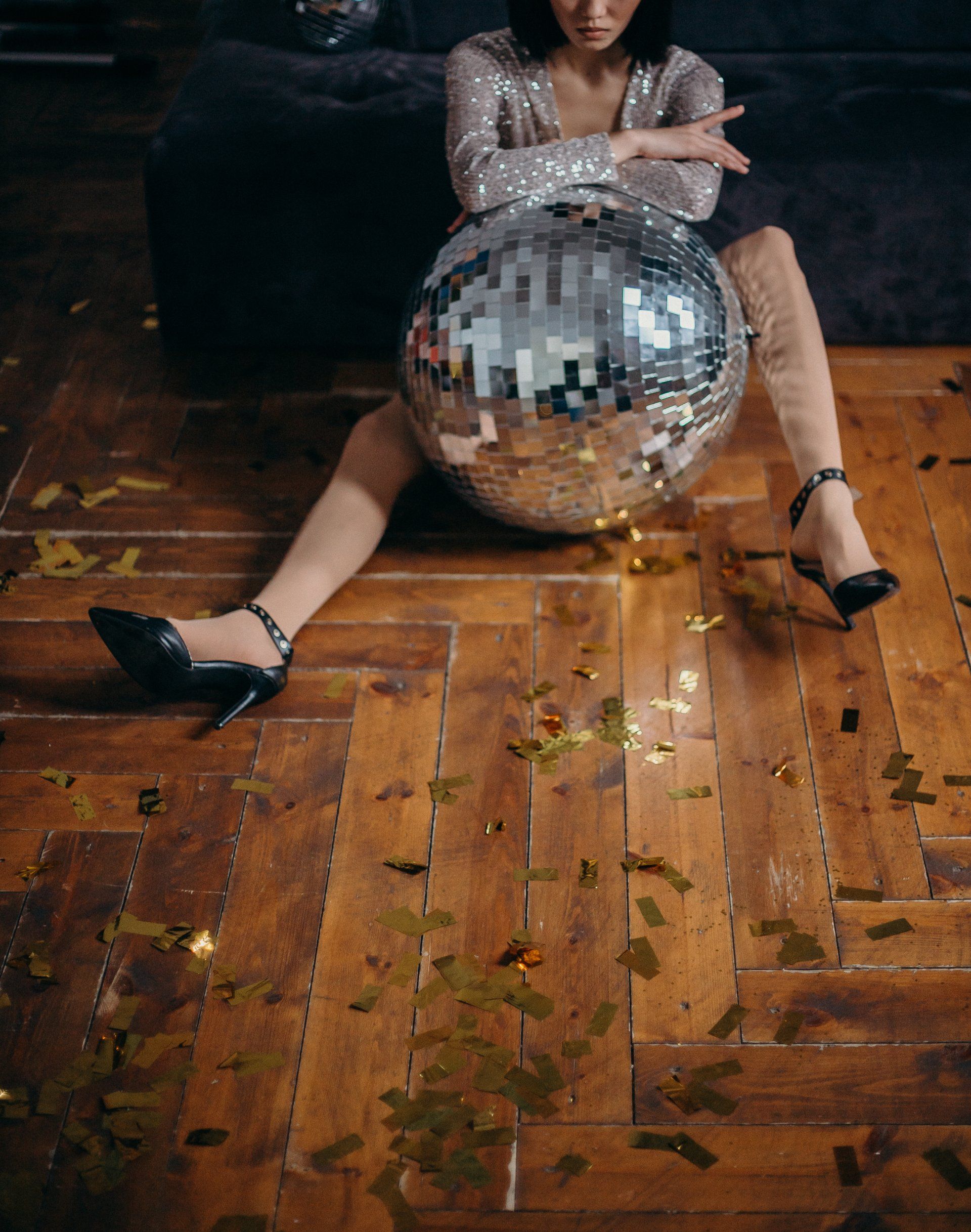 Person in a sparkly dress sitting on a disco ball on a wooden floor, legs splayed.