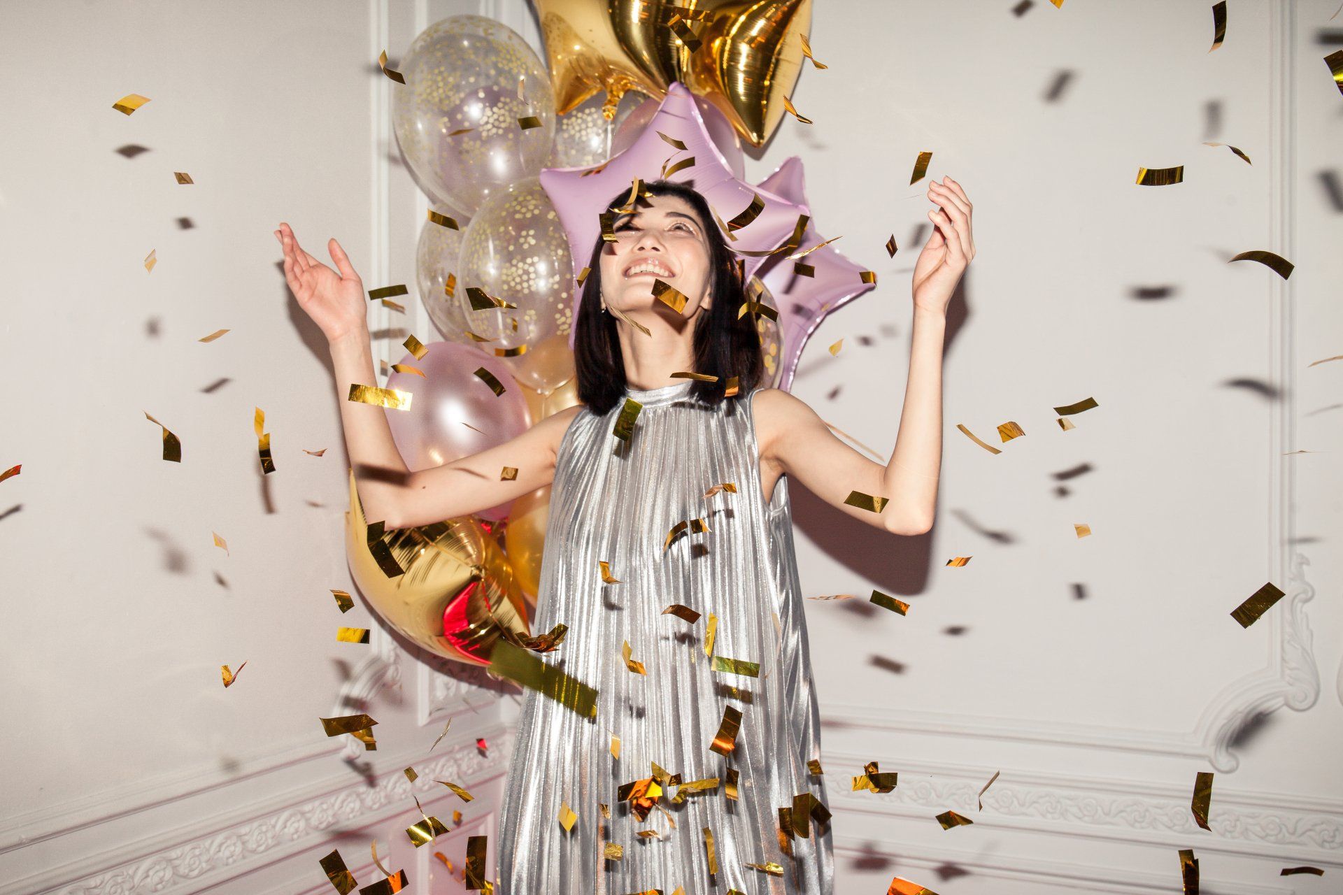 Woman in silver dress throws confetti. Balloons in background.