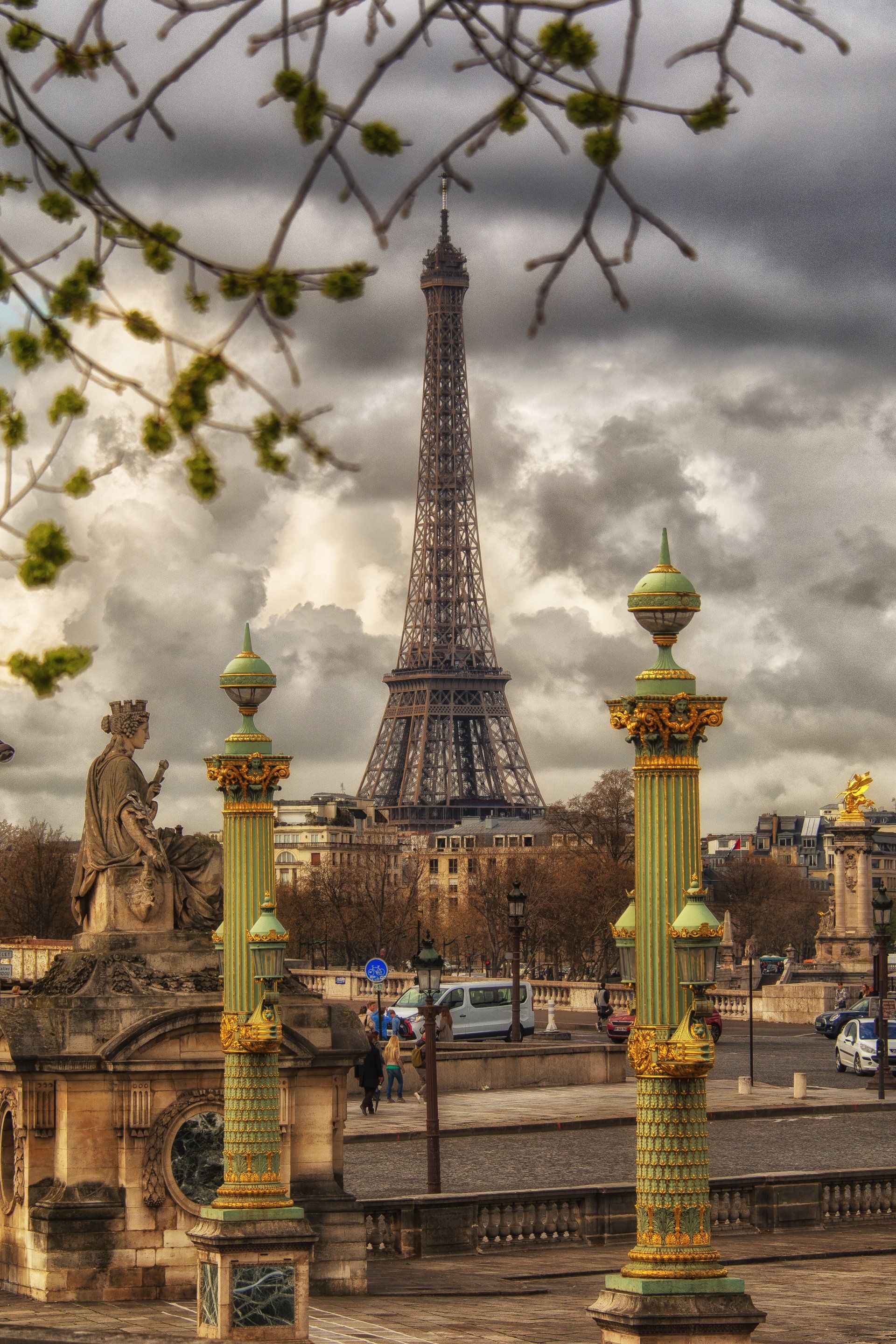 The Eiffel Tower rises behind ornate, gold-trimmed lampposts and a stone statue under a dramatic, cloudy gray sky.