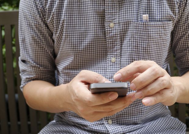 Person holding a smartphone with both hands while seated outdoors, wearing a gray checkered shirt