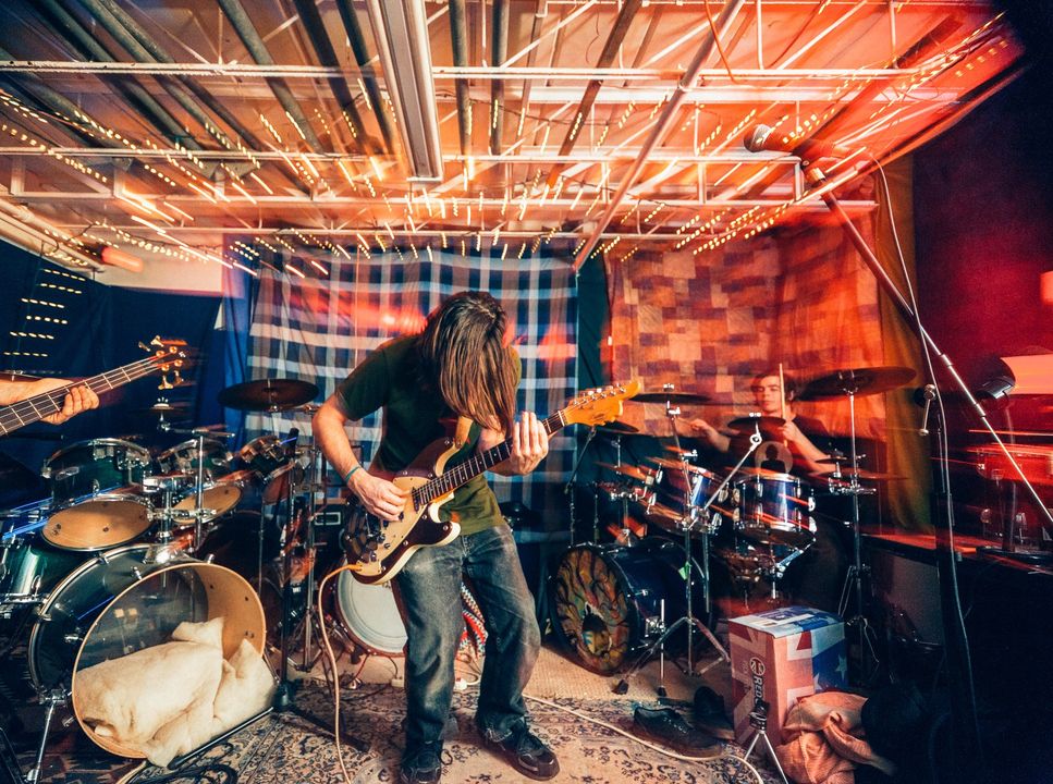 Rock band performing in a small studio, with guitarist center stage under warm orange lighting