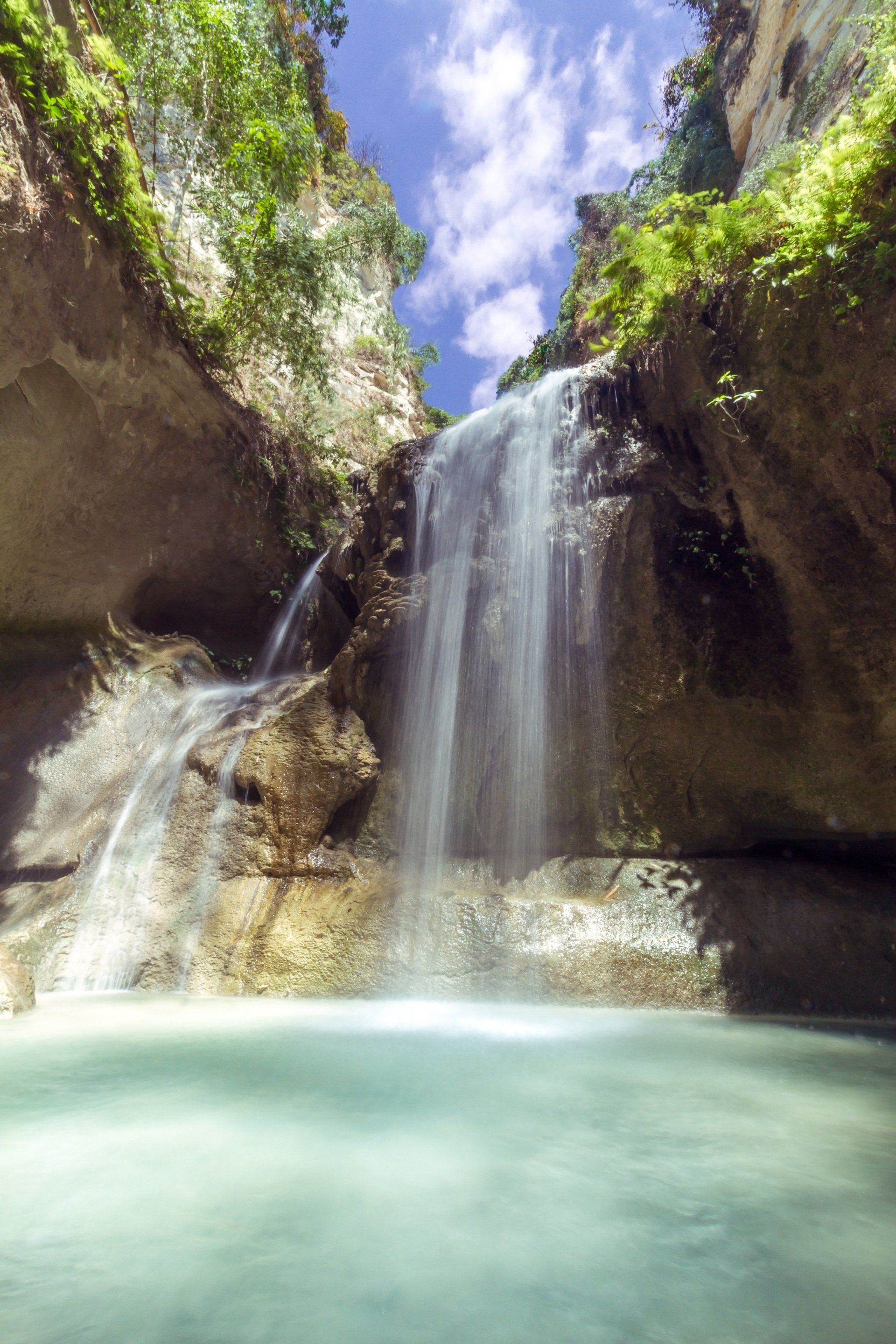 Waterfall cascading into a turquoise pool, surrounded by rock cliffs and lush green foliage under a blue sky.