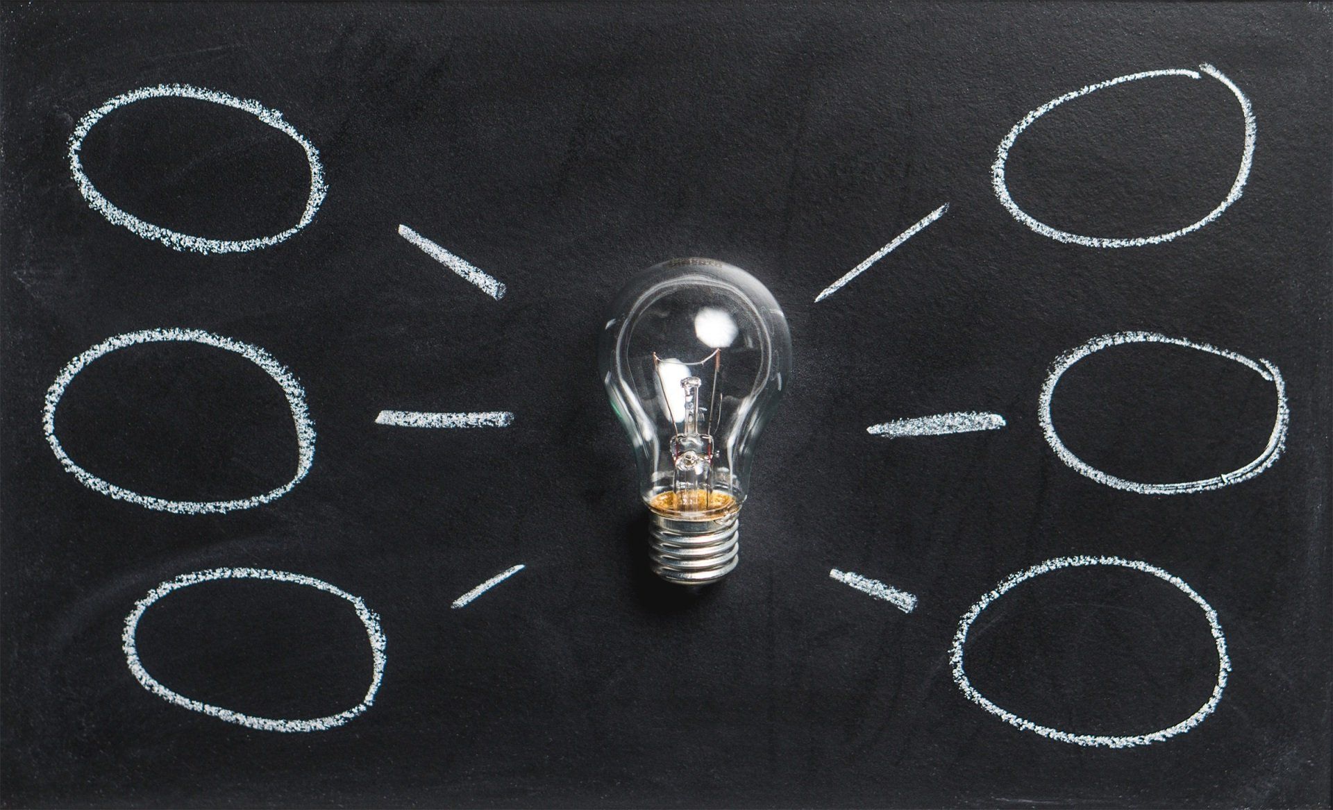 A clear light bulb centered on a chalkboard background, surrounded by six empty chalk-drawn circles connected to the bulb by lines, suggesting a mind map or brainstorming format. The image represents aiarthritis's educational initiatives and awareness programs connecting different aspects of autoinflammatory arthritis knowledge.