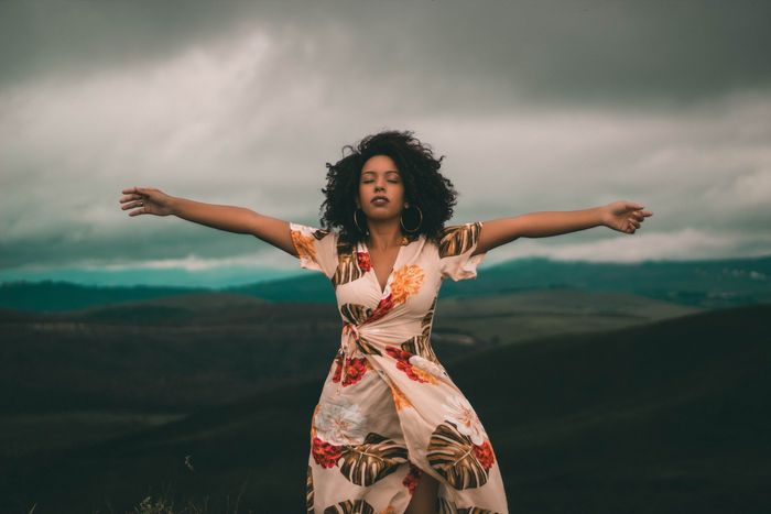 A woman in a floral dress is standing on top of a hill with her arms outstretched.