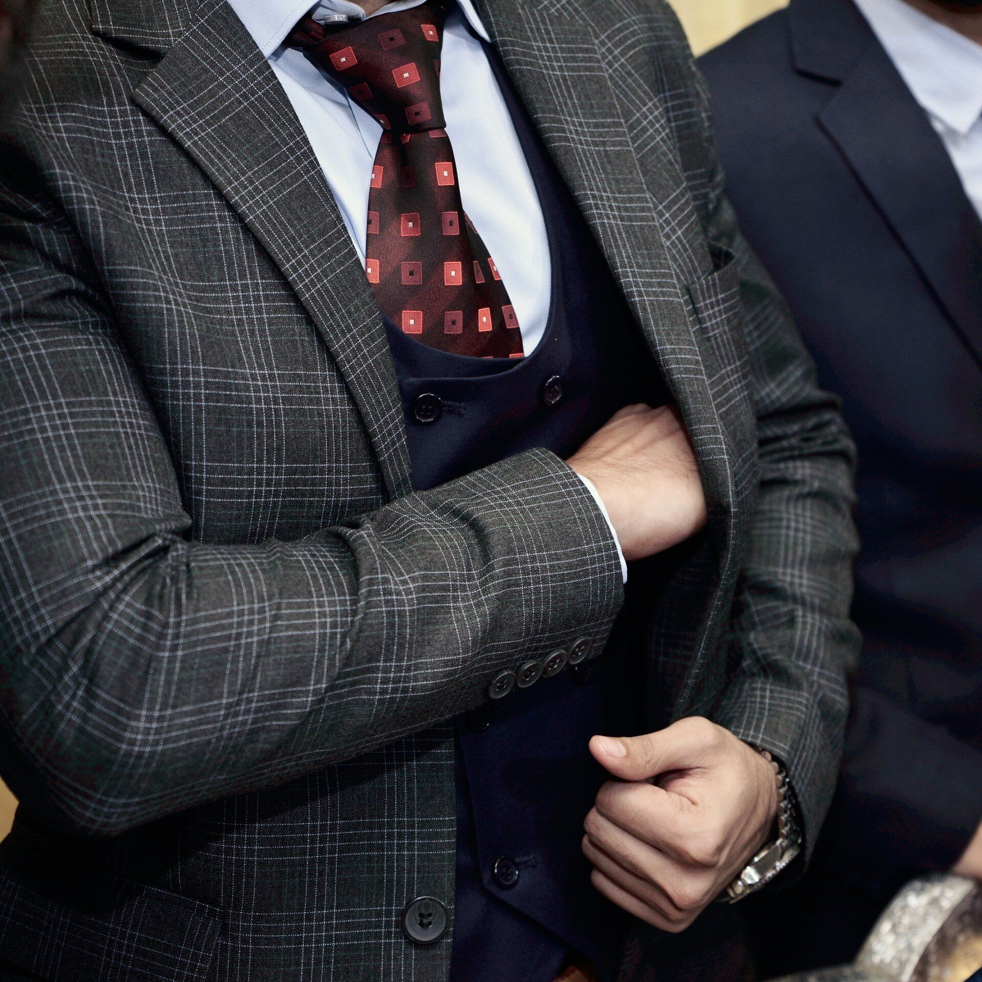 A person in a gray plaid blazer, dark vest, light shirt, and patterned red tie, with one hand inside their jacket pocket.