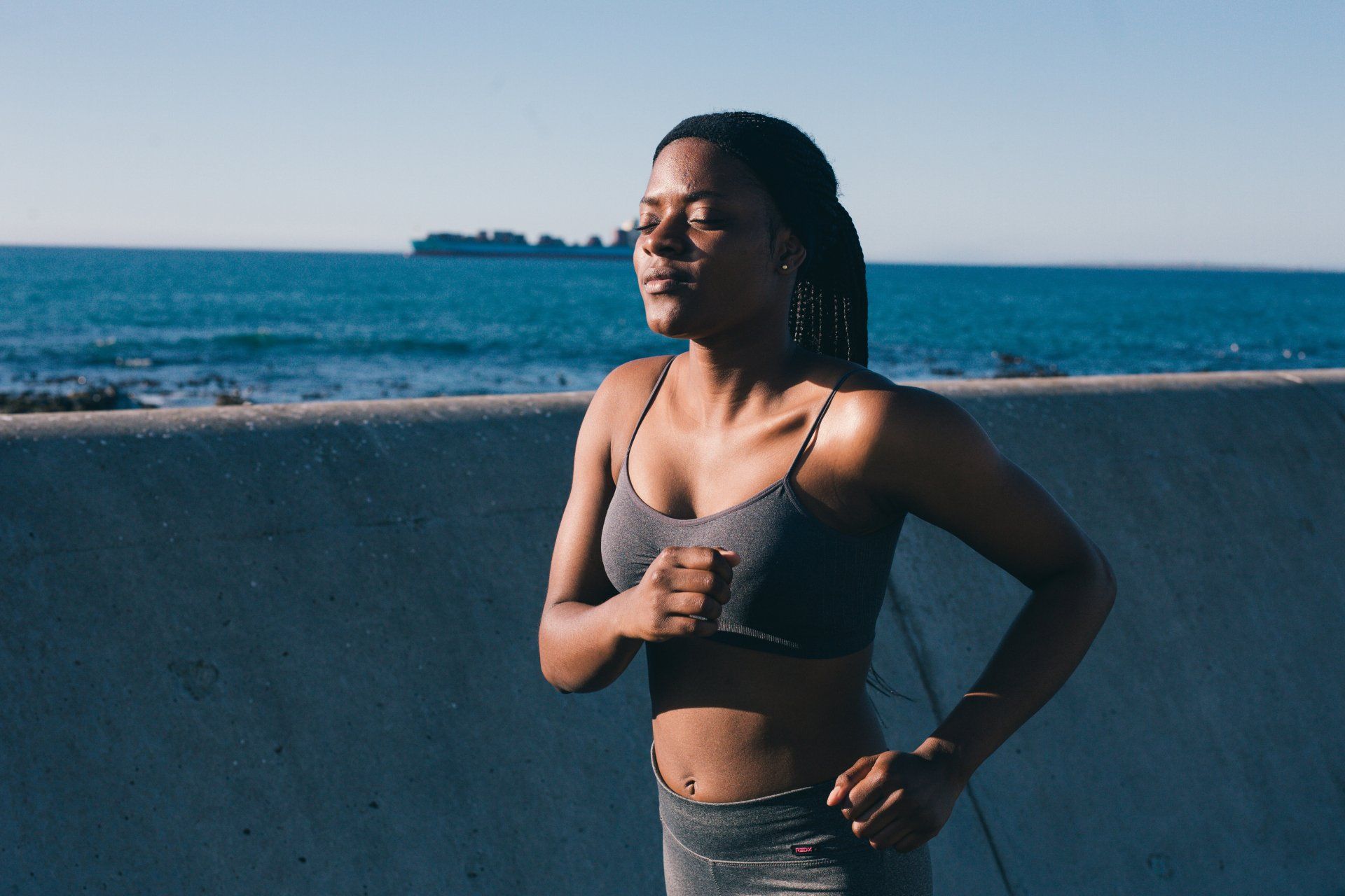 Woman exercising on a seawall with ocean in the background, wearing a sports bra and leggings.