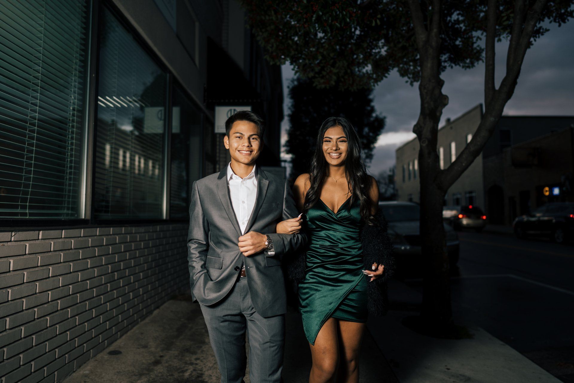 Couple walking arm-in-arm on a sidewalk at dusk. The man wears a suit, and the woman, a green dress.